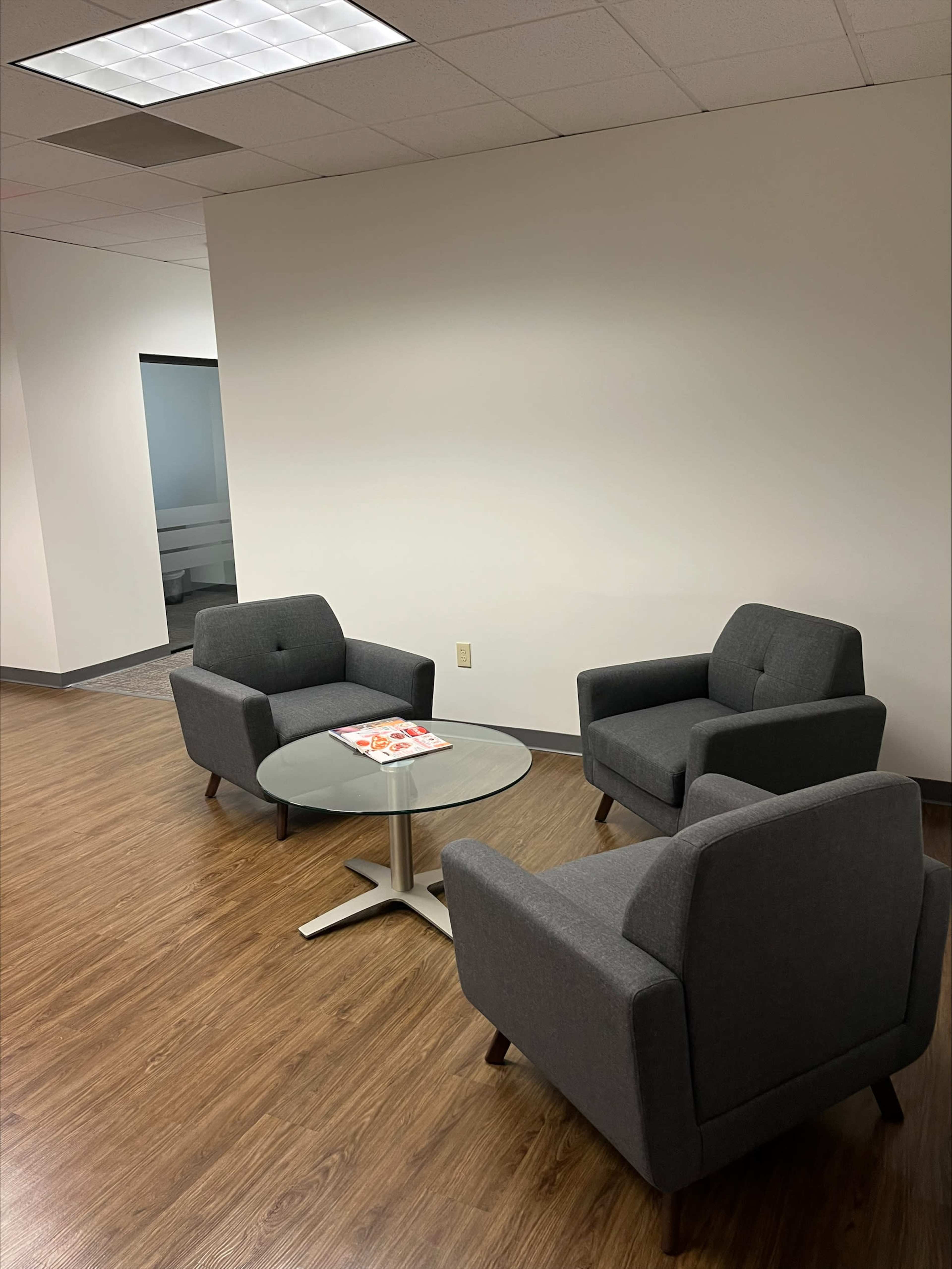 A small seating area with two gray armchairs arranged around a round glass table on a wooden floor.