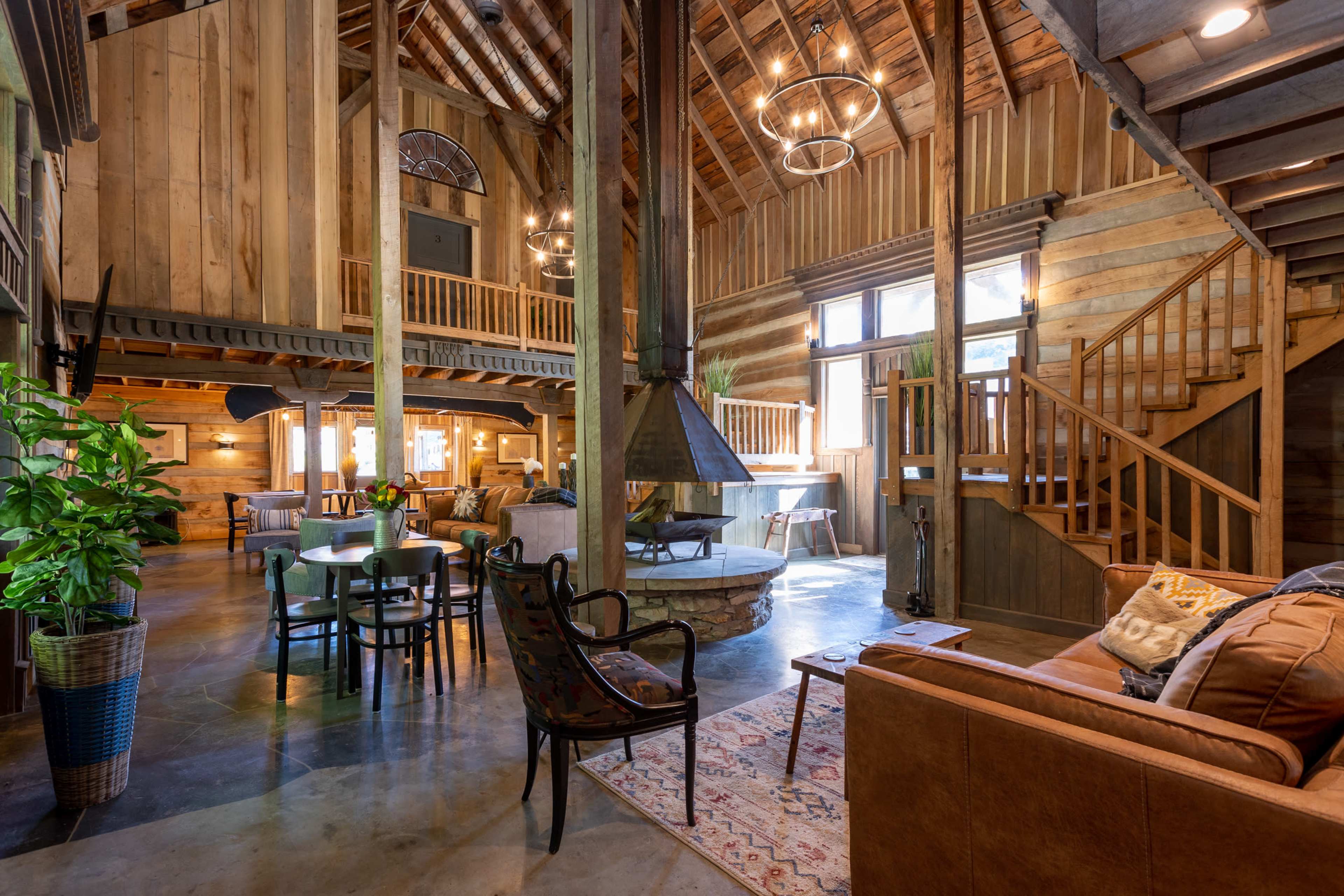 The interior of a rustic cabin features wooden beams, a stone fireplace, and a cozy seating area with a staircase leading to an upper level.