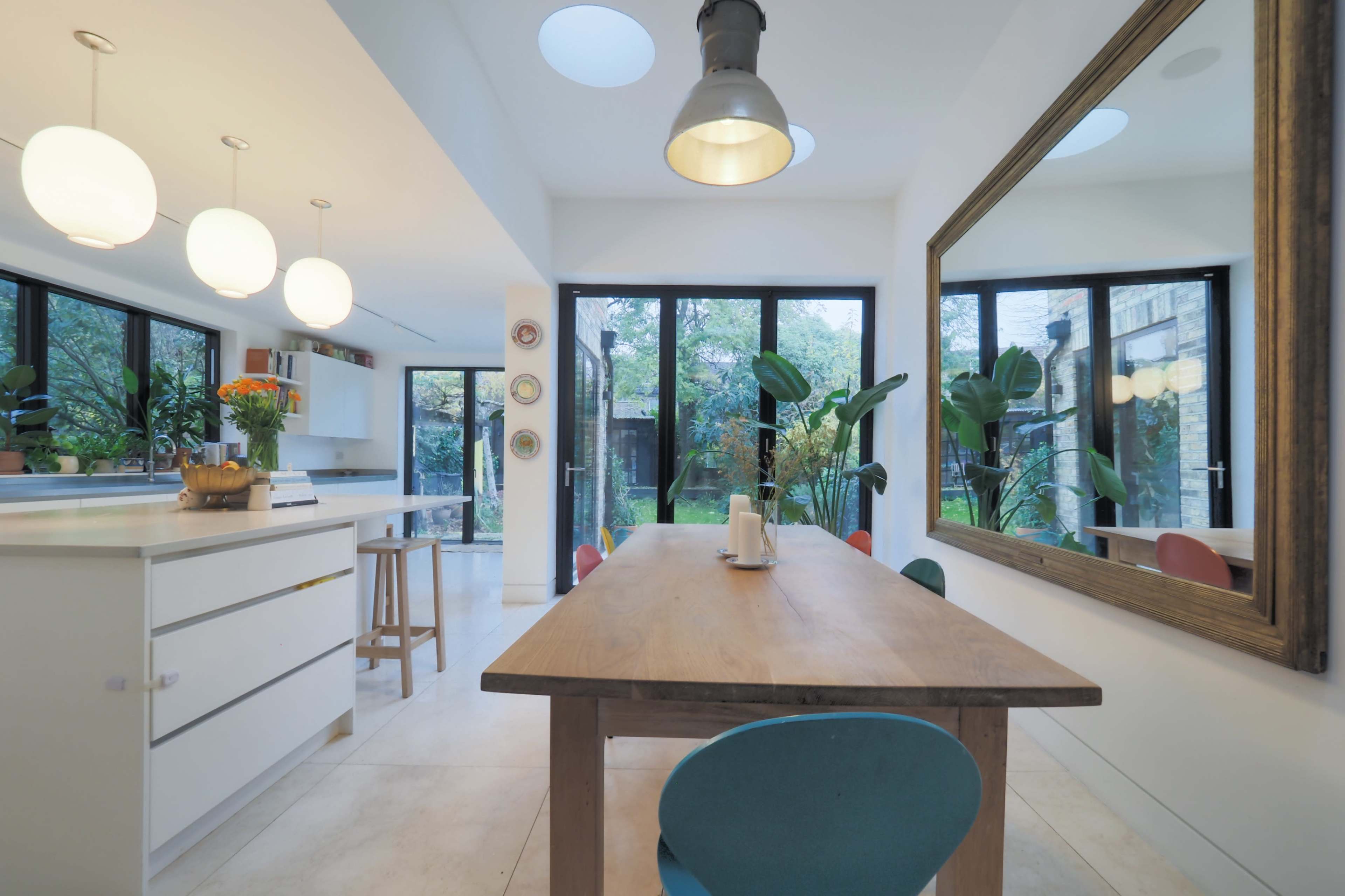 The image shows a modern kitchen and dining area with a wooden table, pendant lighting, and large glass windows overlooking a garden.