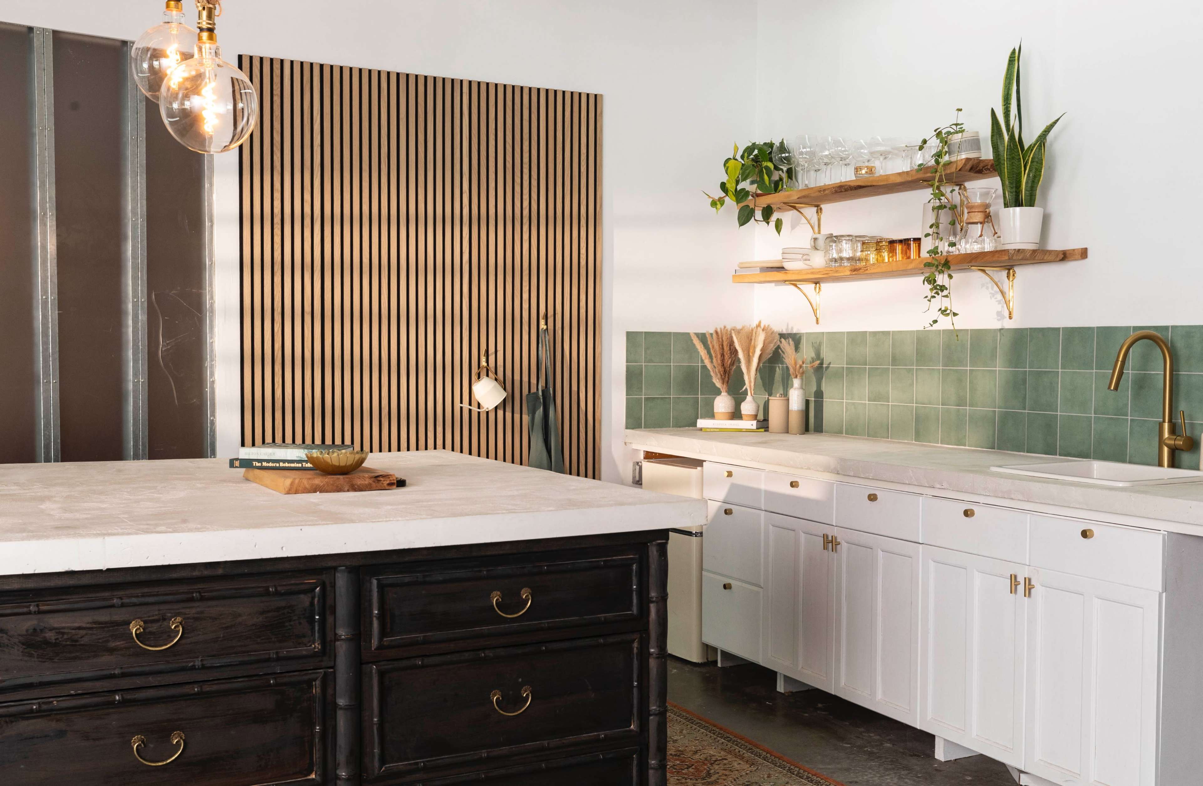 A modern kitchen with a light-colored island, white cabinetry, and green tiled backsplash, complemented by wooden shelves adorned with plants and decorative items.