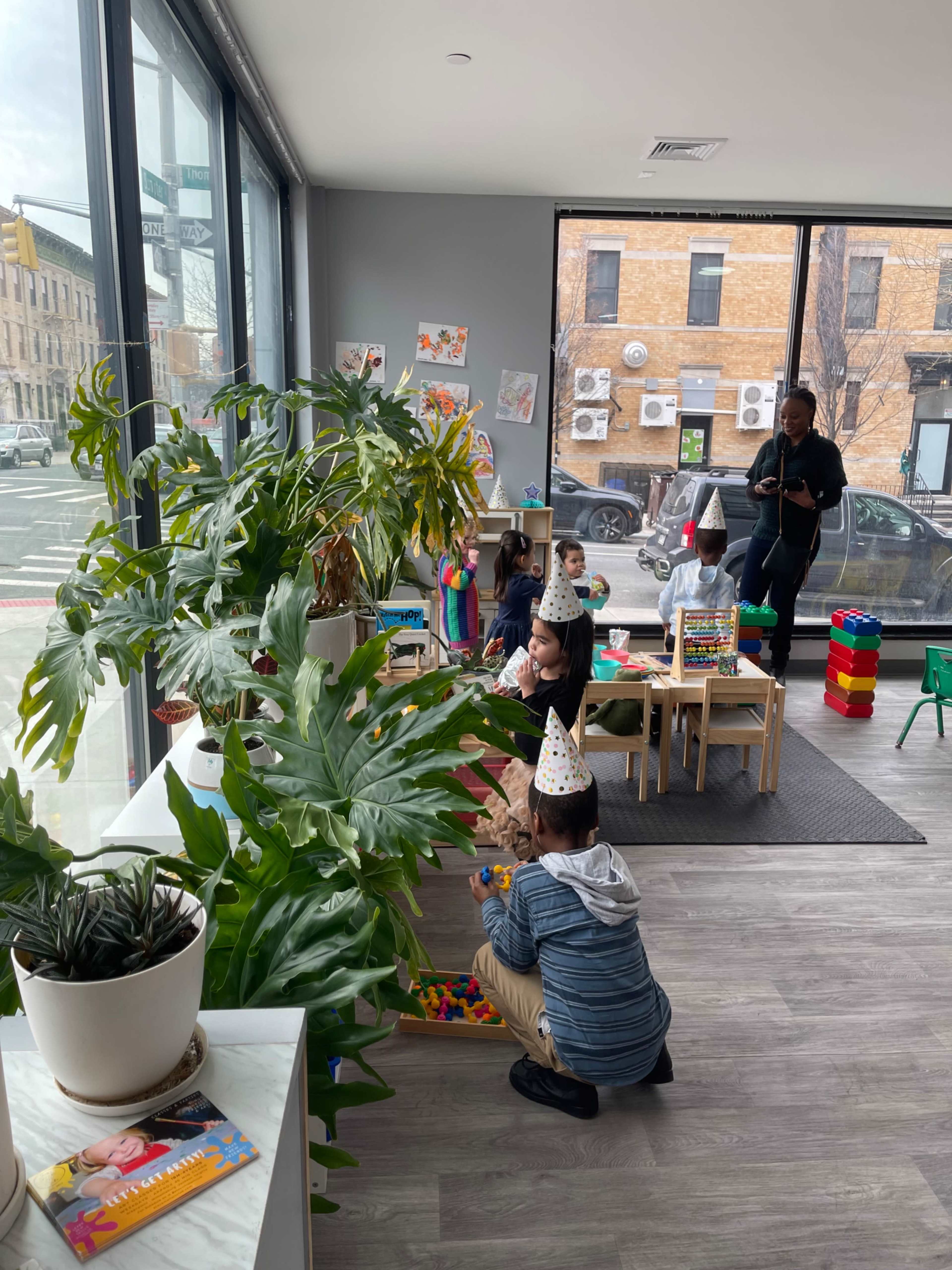 A group of children wearing party hats play with building blocks in a brightly lit room, while an adult supervises from the background.