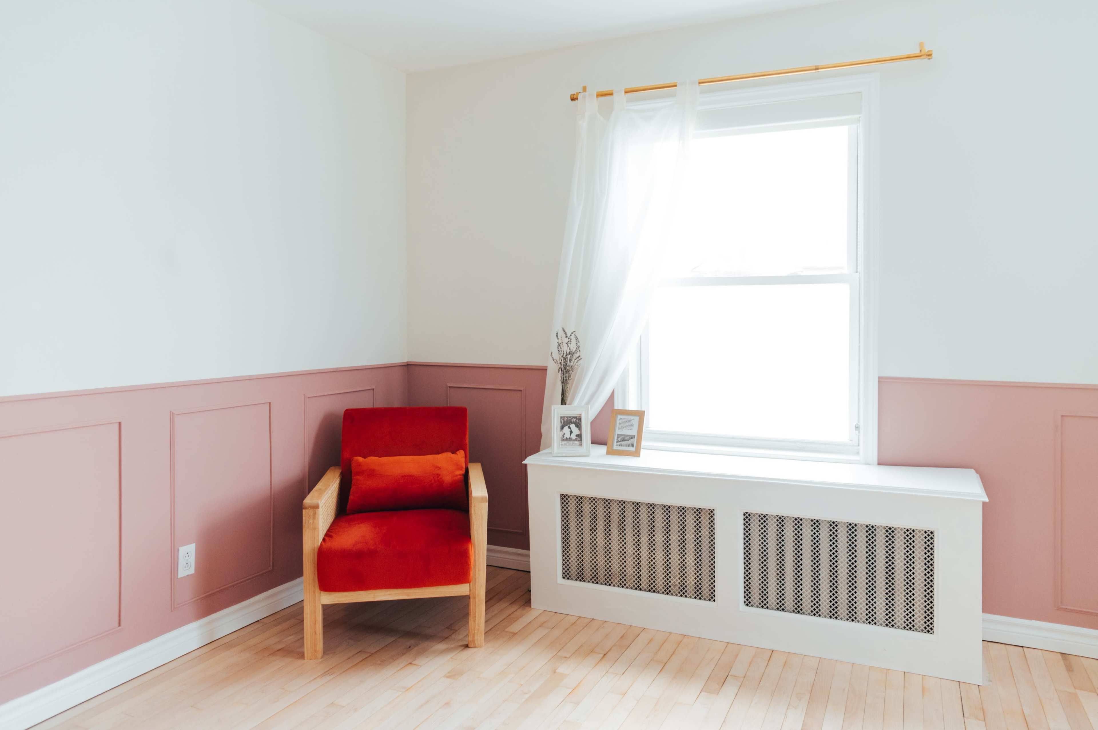 A bright, minimalist room with a red upholstered chair beside a window dressed with sheer curtains and a white radiator under it.