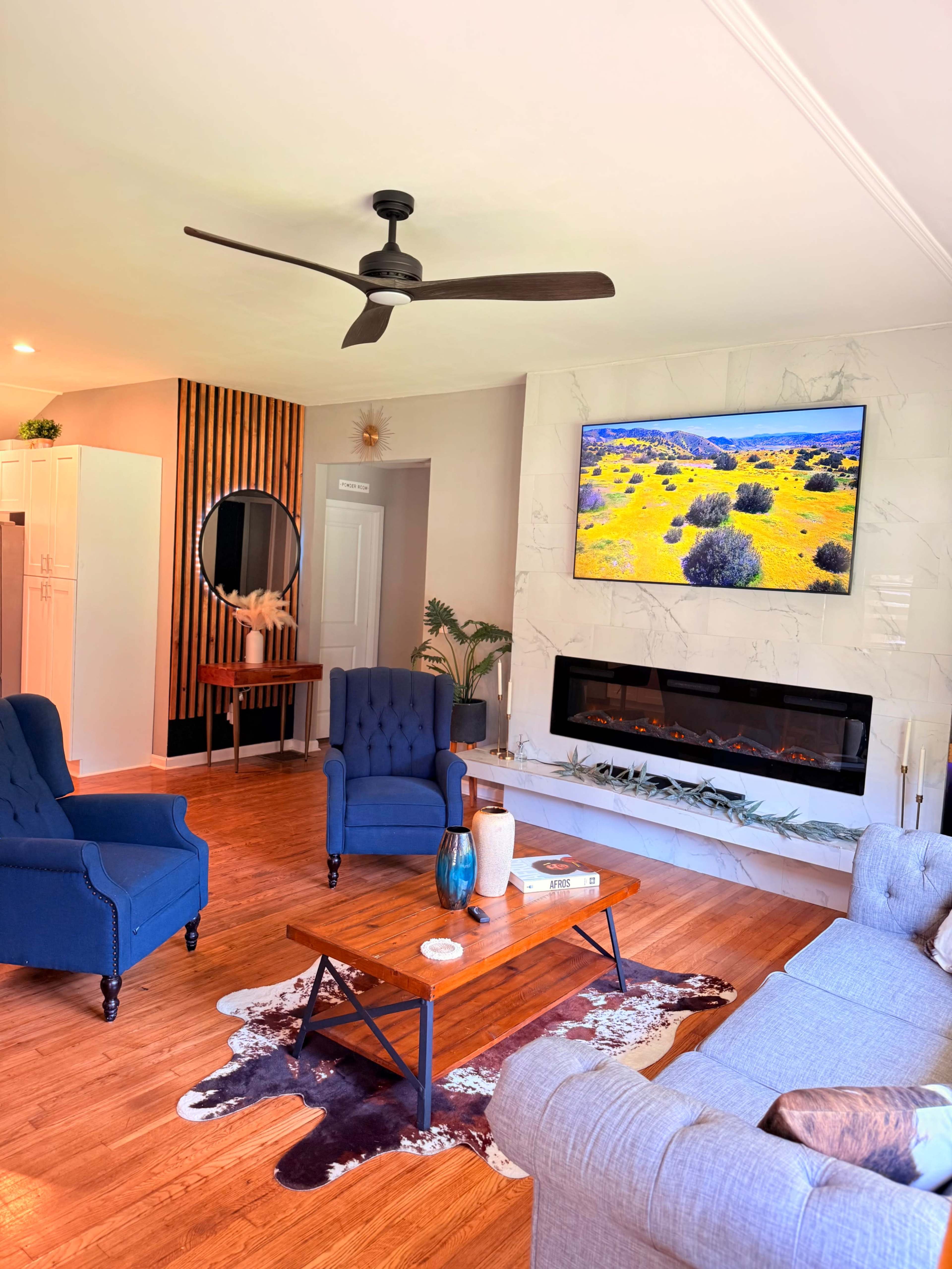 The living room features a TV mounted above a modern fireplace, surrounded by two blue armchairs, a gray sofa, and a wooden coffee table on a decorative rug.