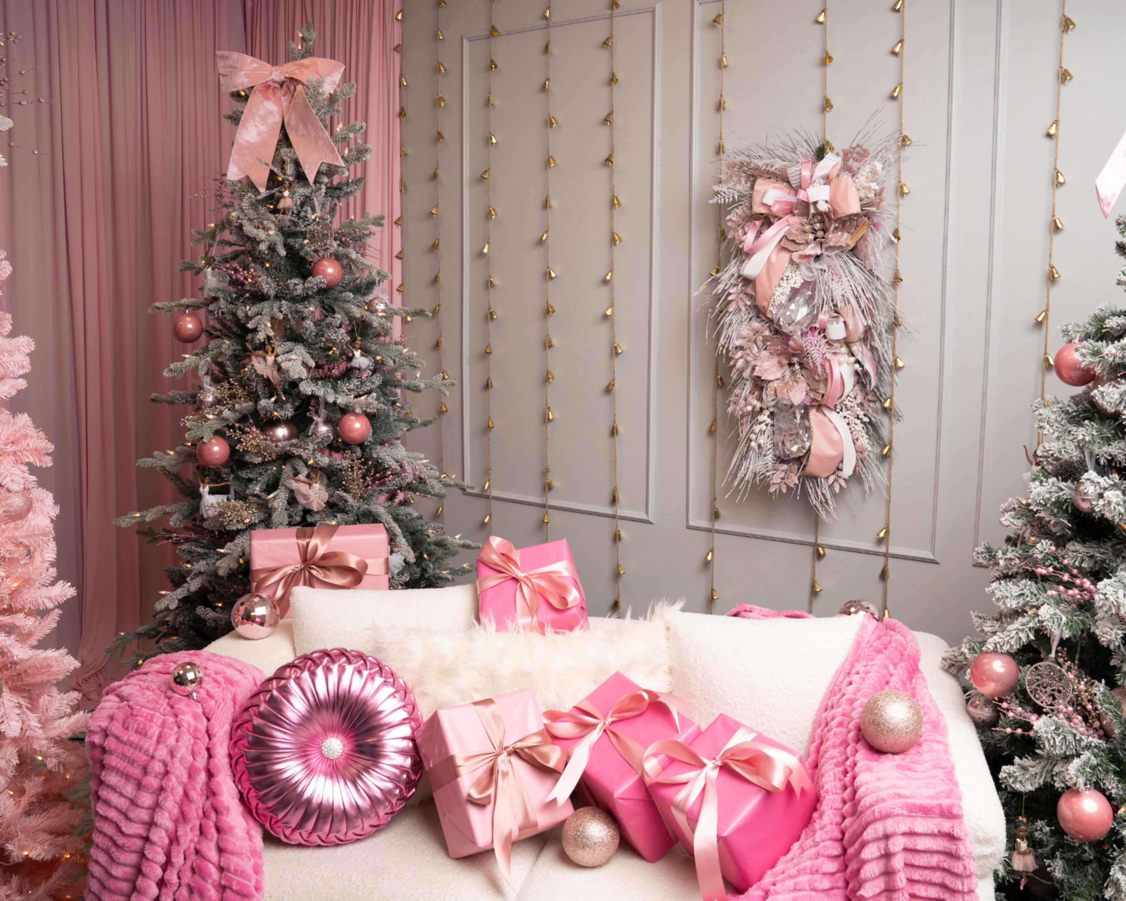 The image features a festive room decorated with pink Christmas trees, pink-wrapped gifts, and a wreath on the wall, all set against a backdrop of soft pink curtains and wall decor.