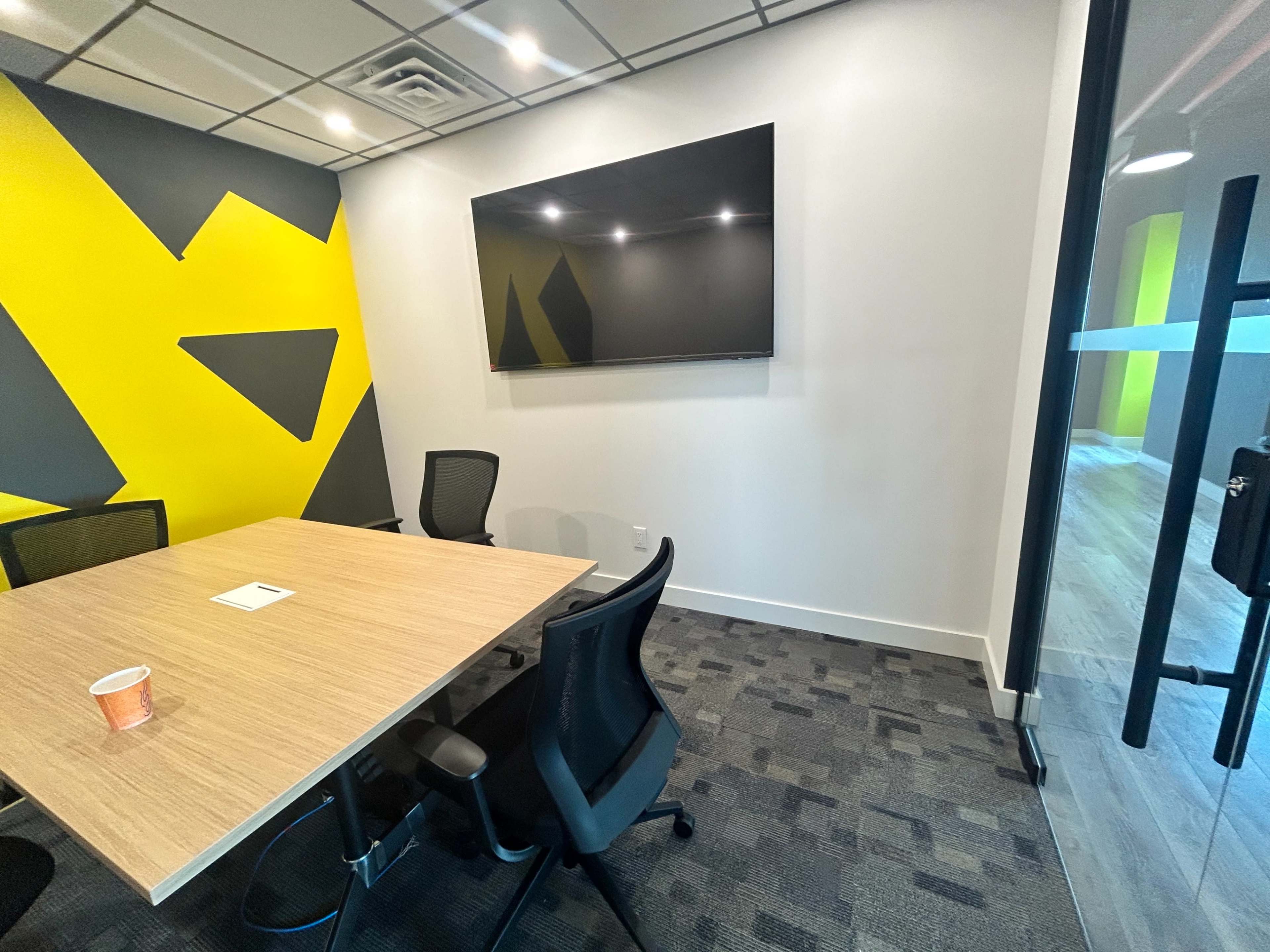 A modern conference room features a wooden table surrounded by black chairs and a wall adorned with yellow and black geometric patterns, with a large screen mounted on one side.