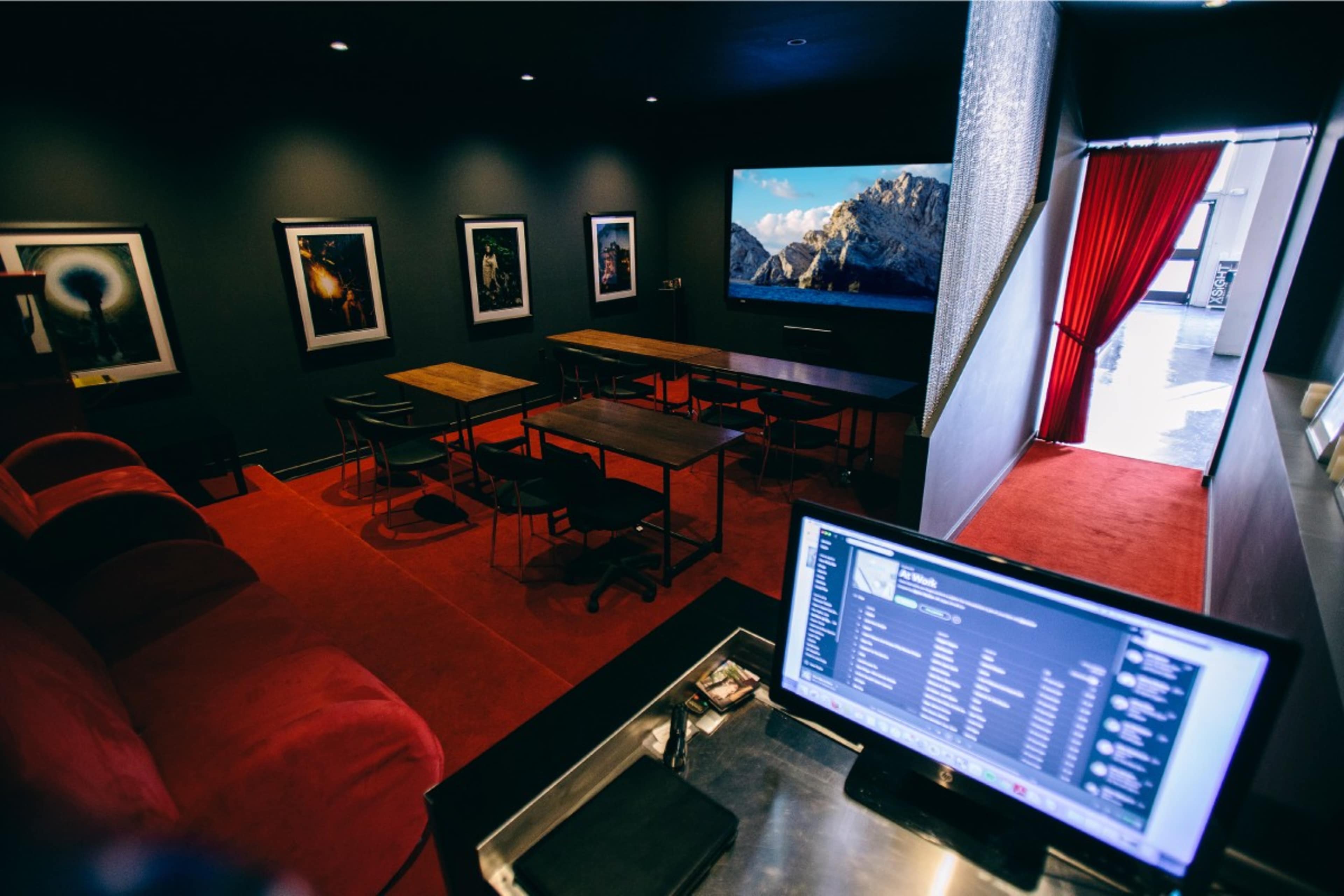 A dimly lit room with a red carpet, featuring a large screen, seating areas, and tables arranged for meetings or presentations.