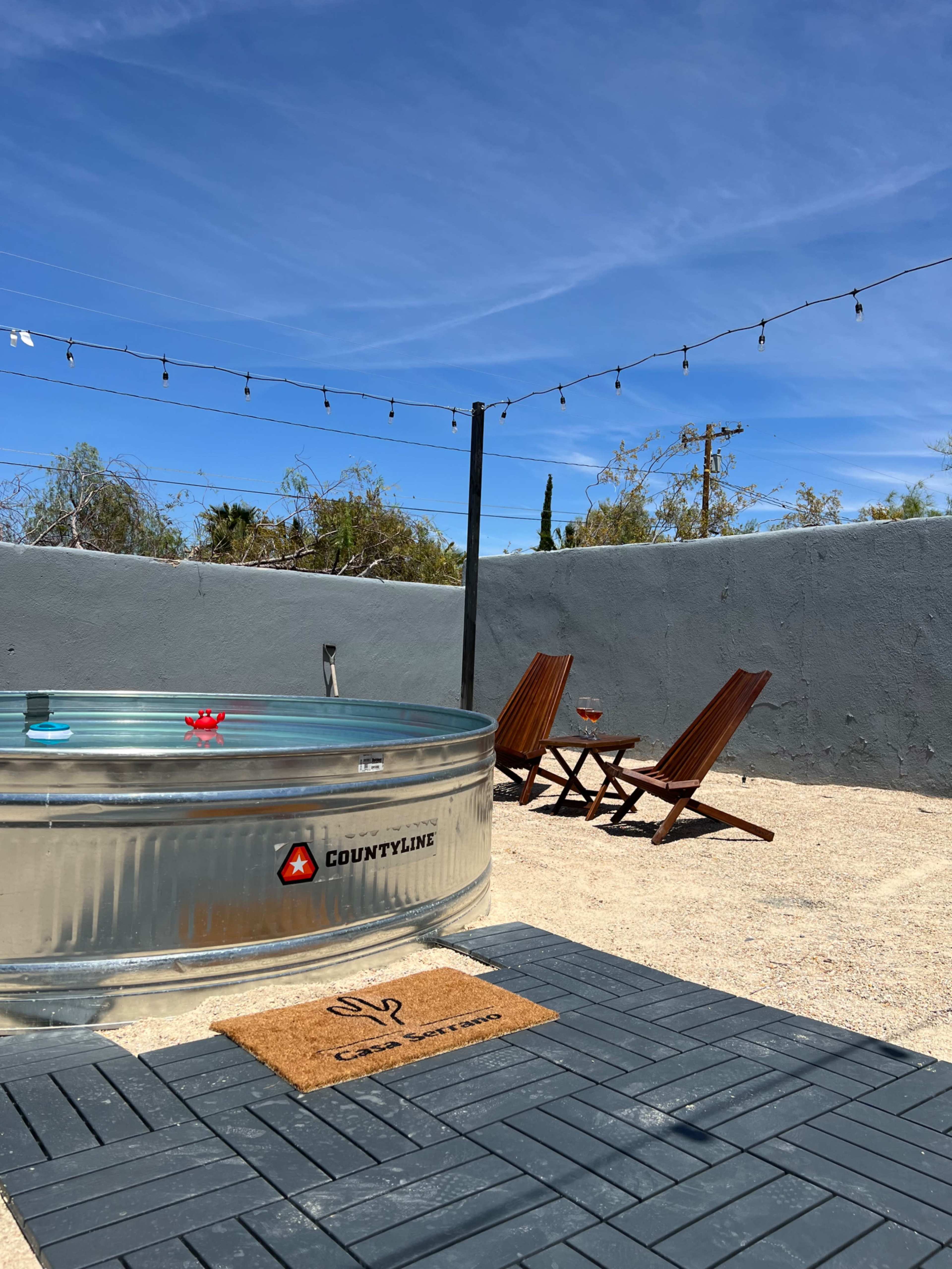 A metal hot tub sits on a gravel patio beside two wooden lounge chairs under string lights.