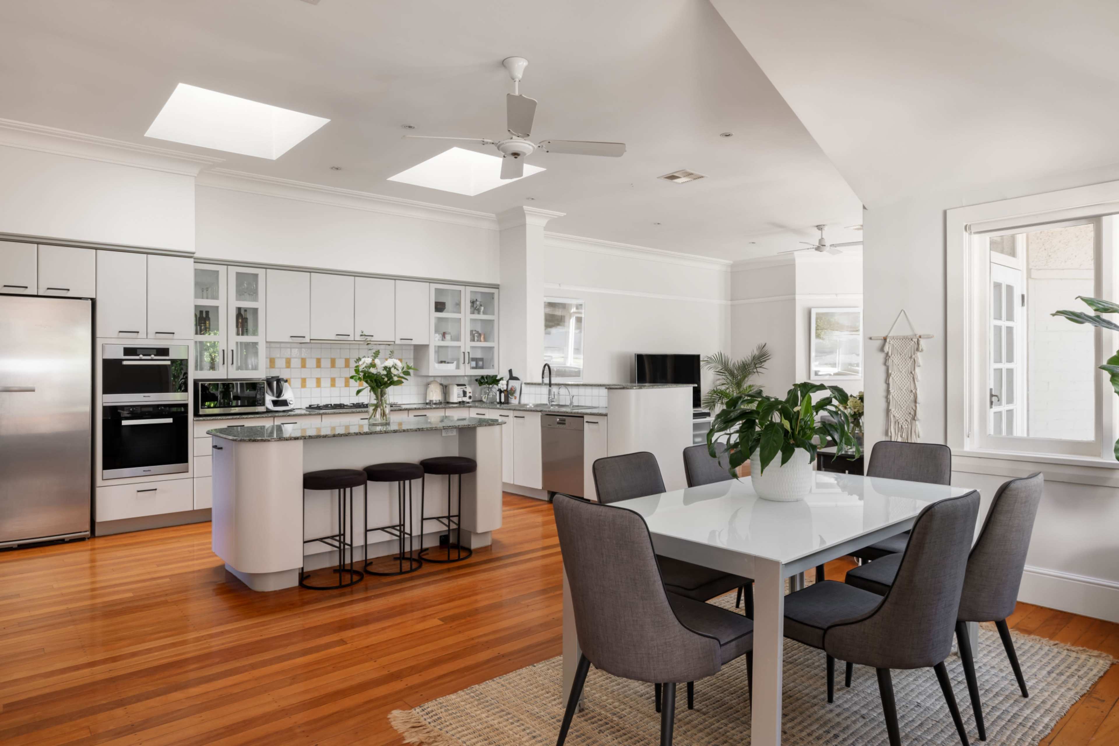 A modern kitchen features a central island with bar stools, a dining table, and large windows allowing natural light.