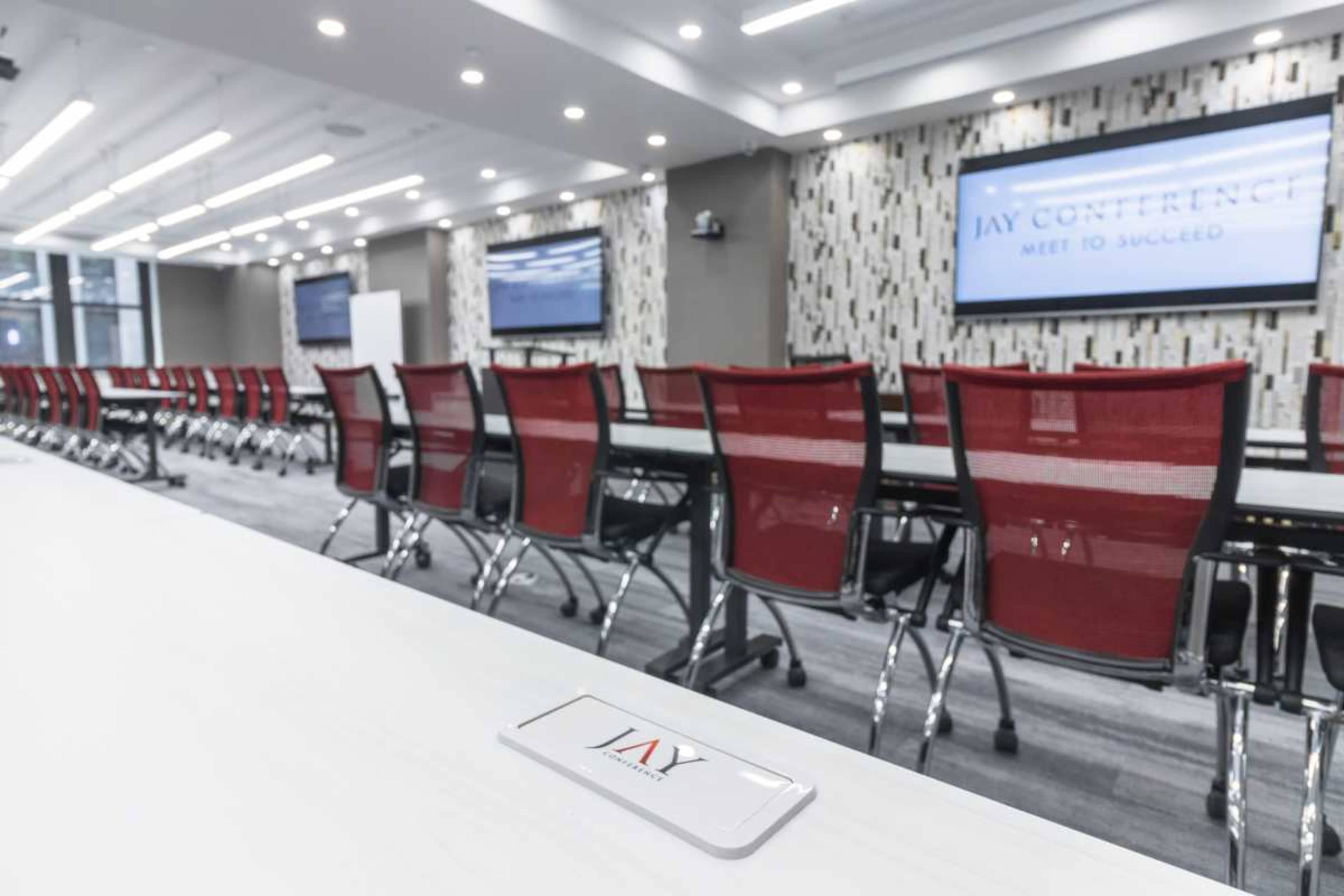 The image shows a modern conference room with red mesh chairs arranged around long white tables and large screens on the walls.