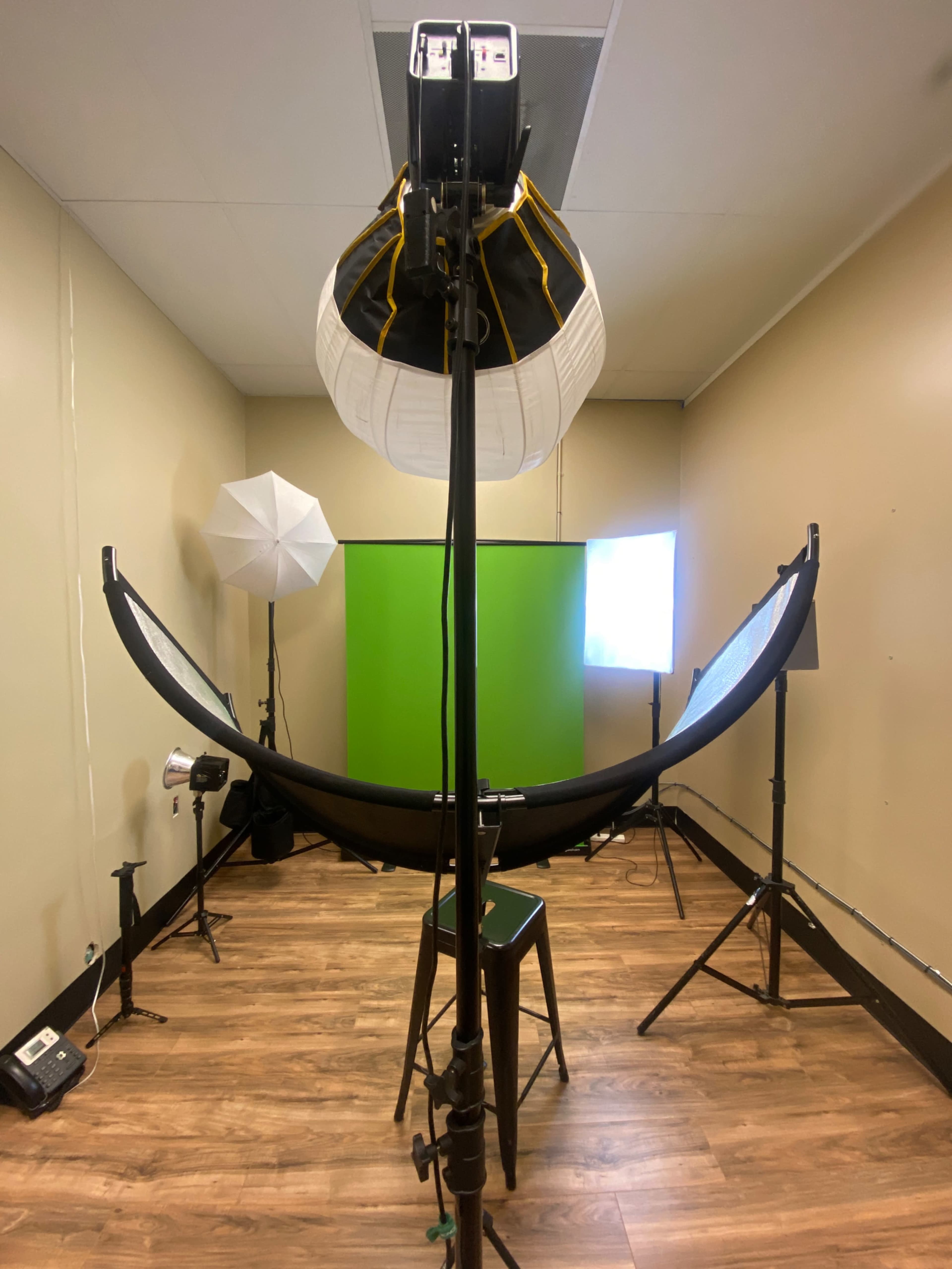 A studio setup featuring a green backdrop, various lighting equipment, and a stool in a room with wood flooring.