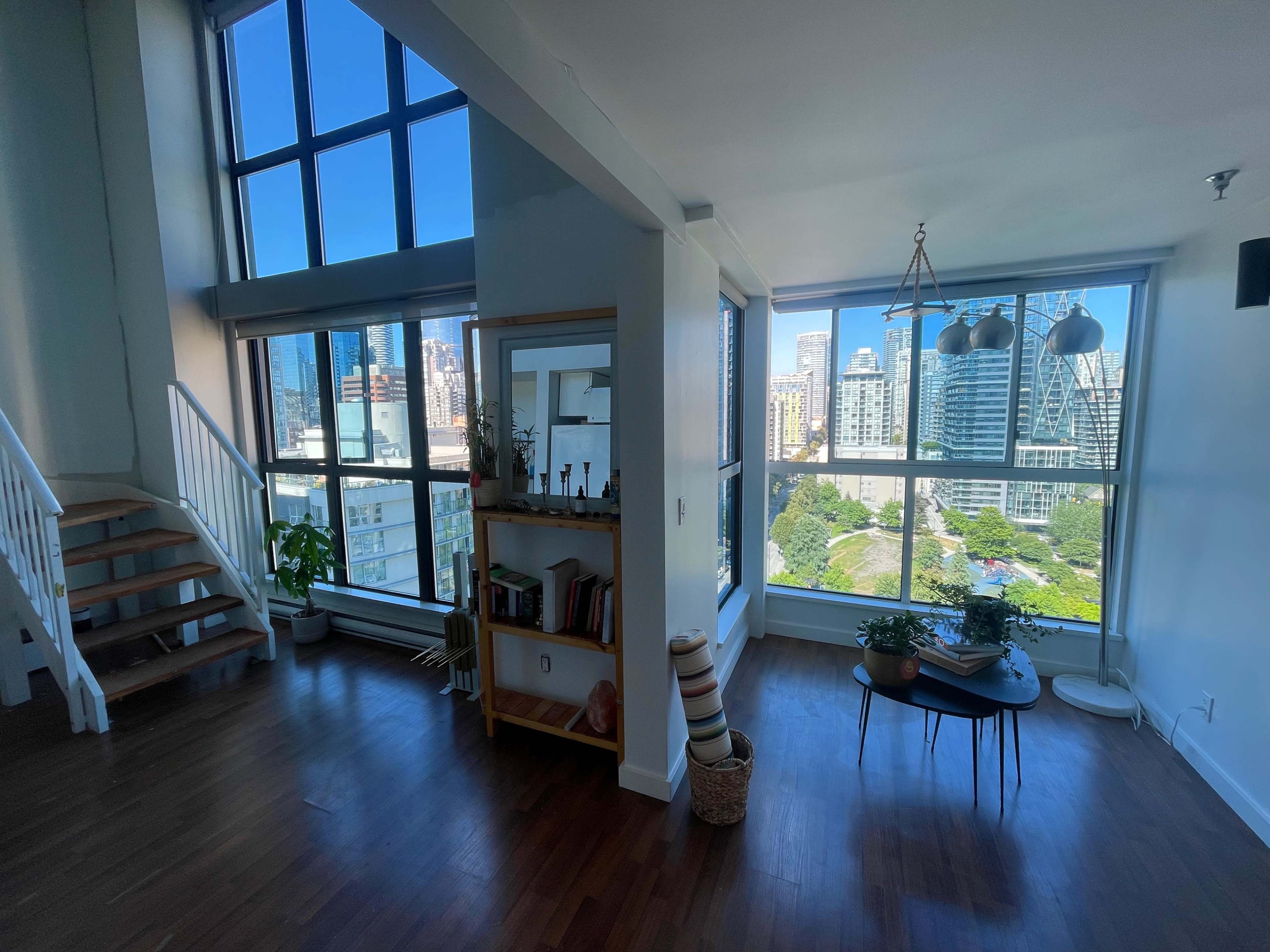 A bright, spacious living area features large windows with city views, a staircase, a bookshelf, and a small round table.