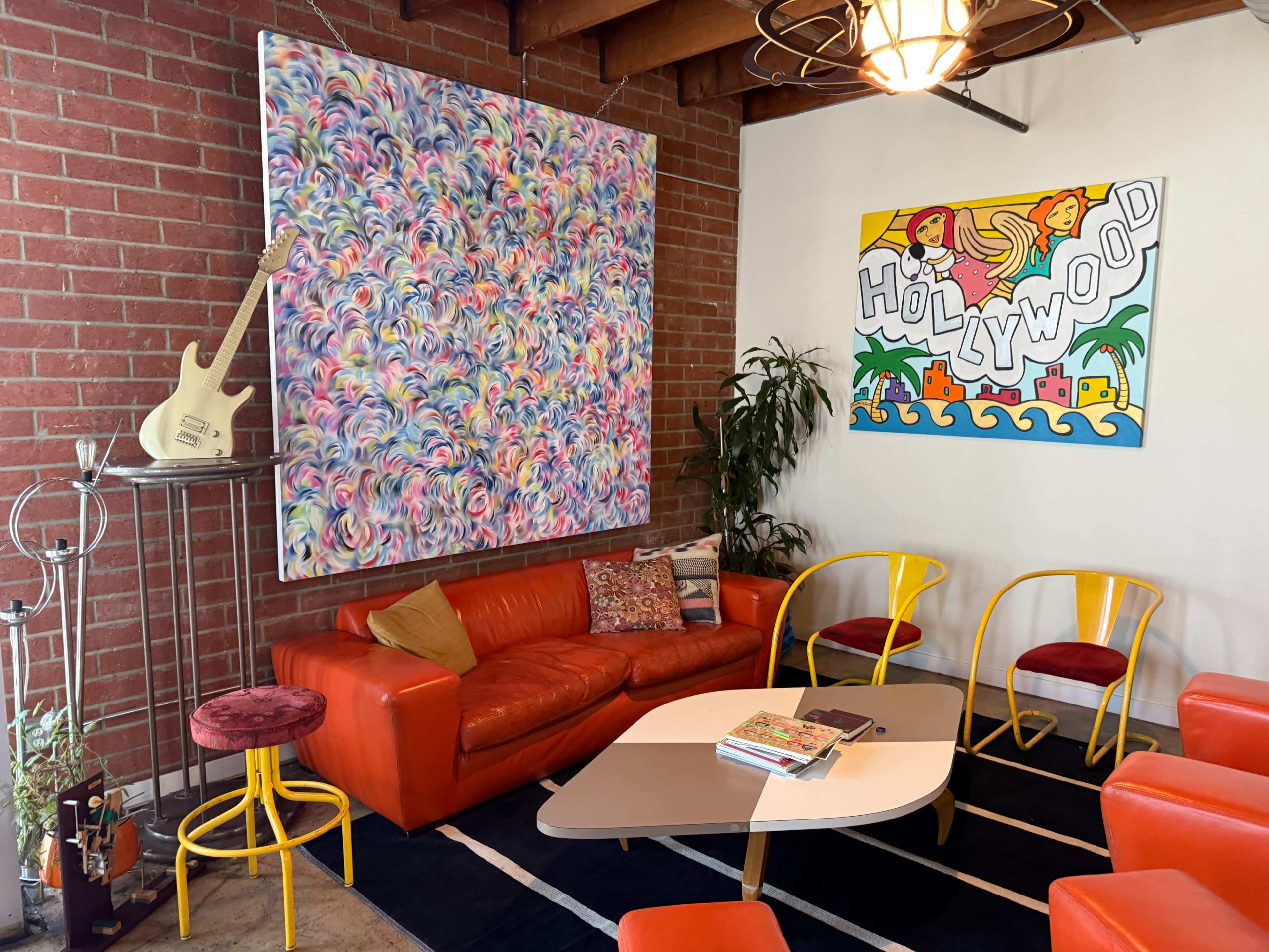 A vibrant living space features a colorful abstract painting, a large red couch, yellow chairs, and a coffee table on a black rug against a backdrop of exposed brick walls.