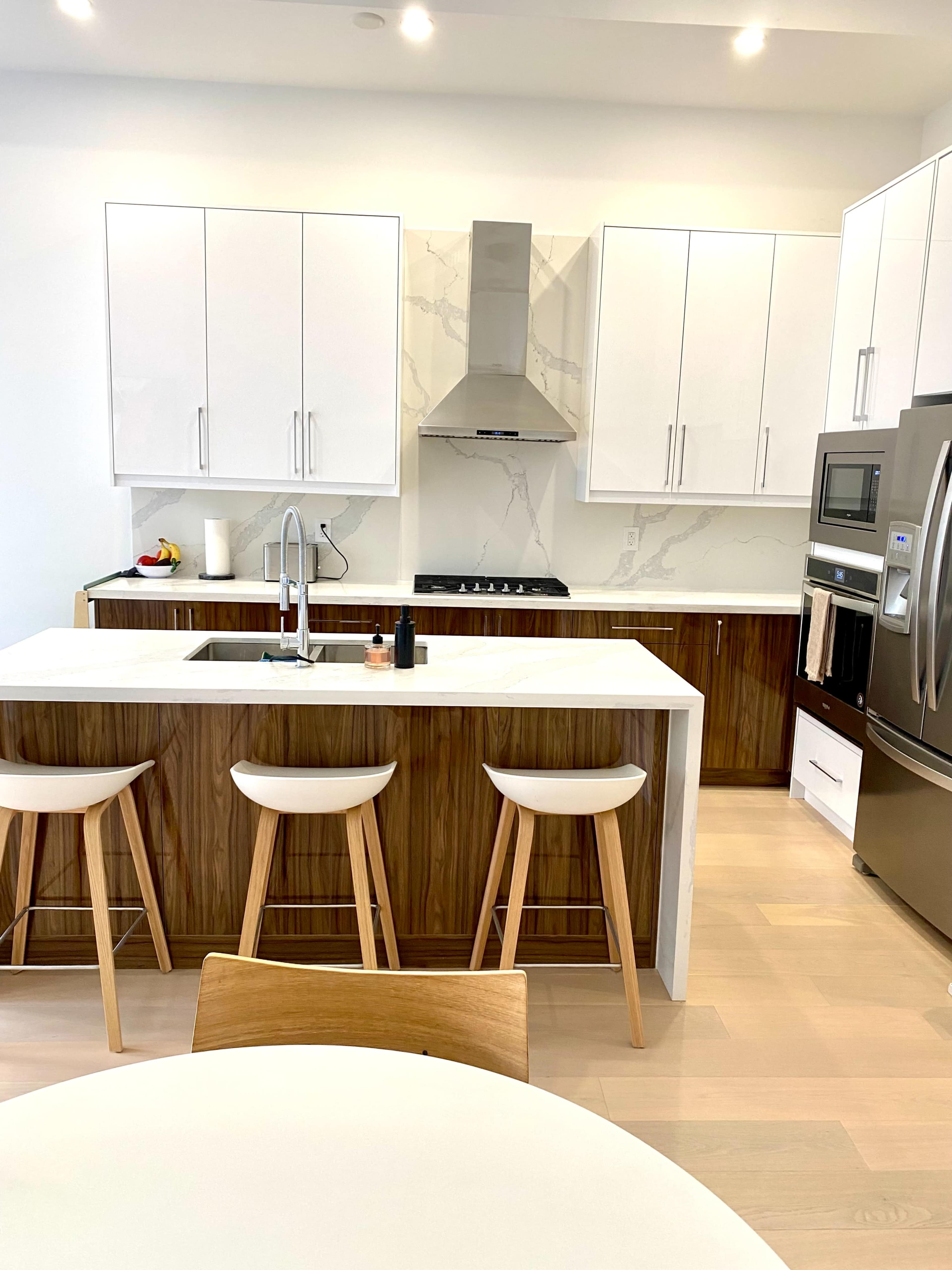 A modern kitchen features white cabinetry, a marble backsplash, and stainless steel appliances, with a central island and three wooden stools.