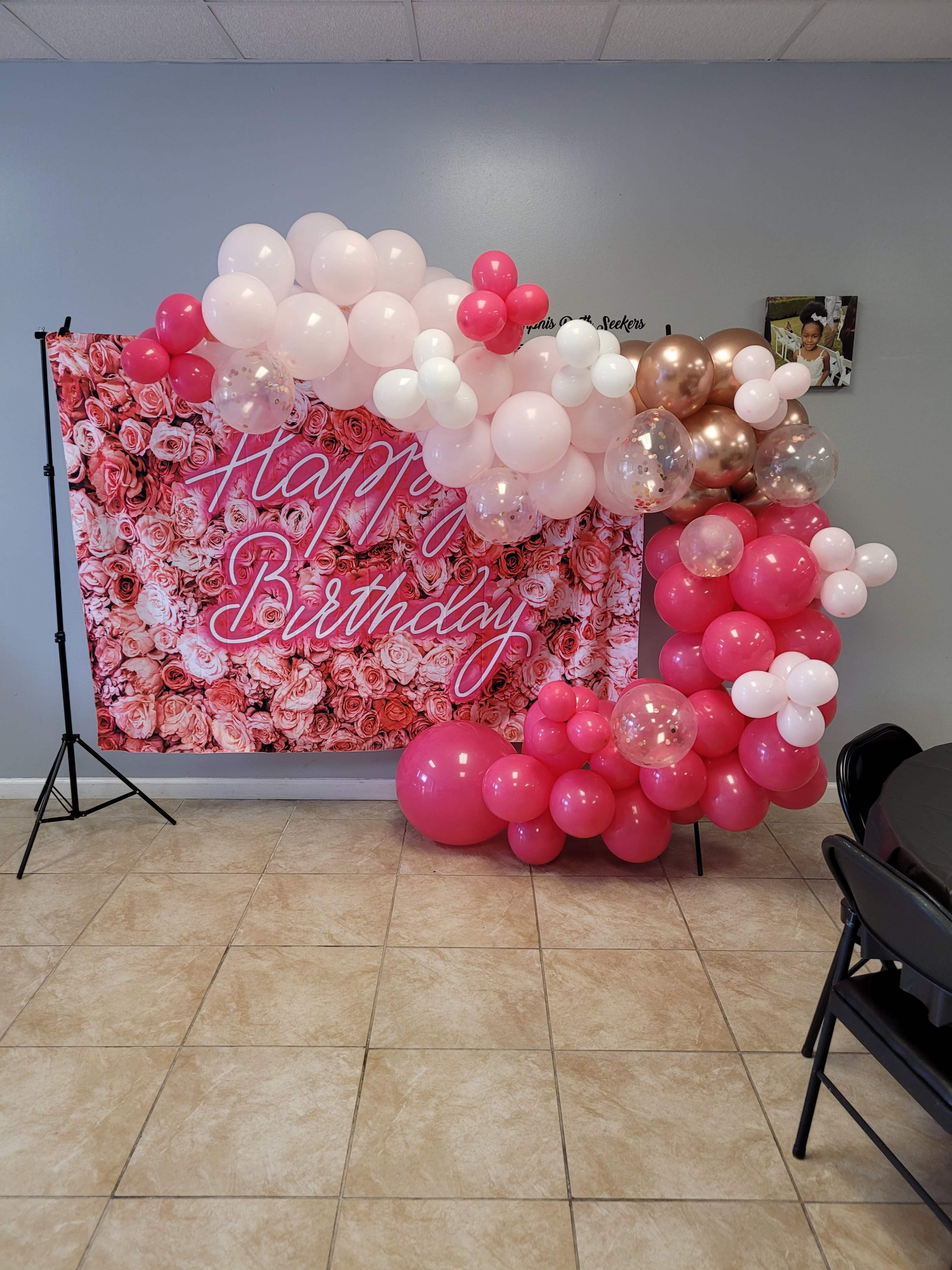 The image shows a birthday decoration setup featuring a backdrop of pink roses with the words "Happy Birthday" in bold letters, accompanied by a colorful arrangement of balloons in various shades of pink and gold.
