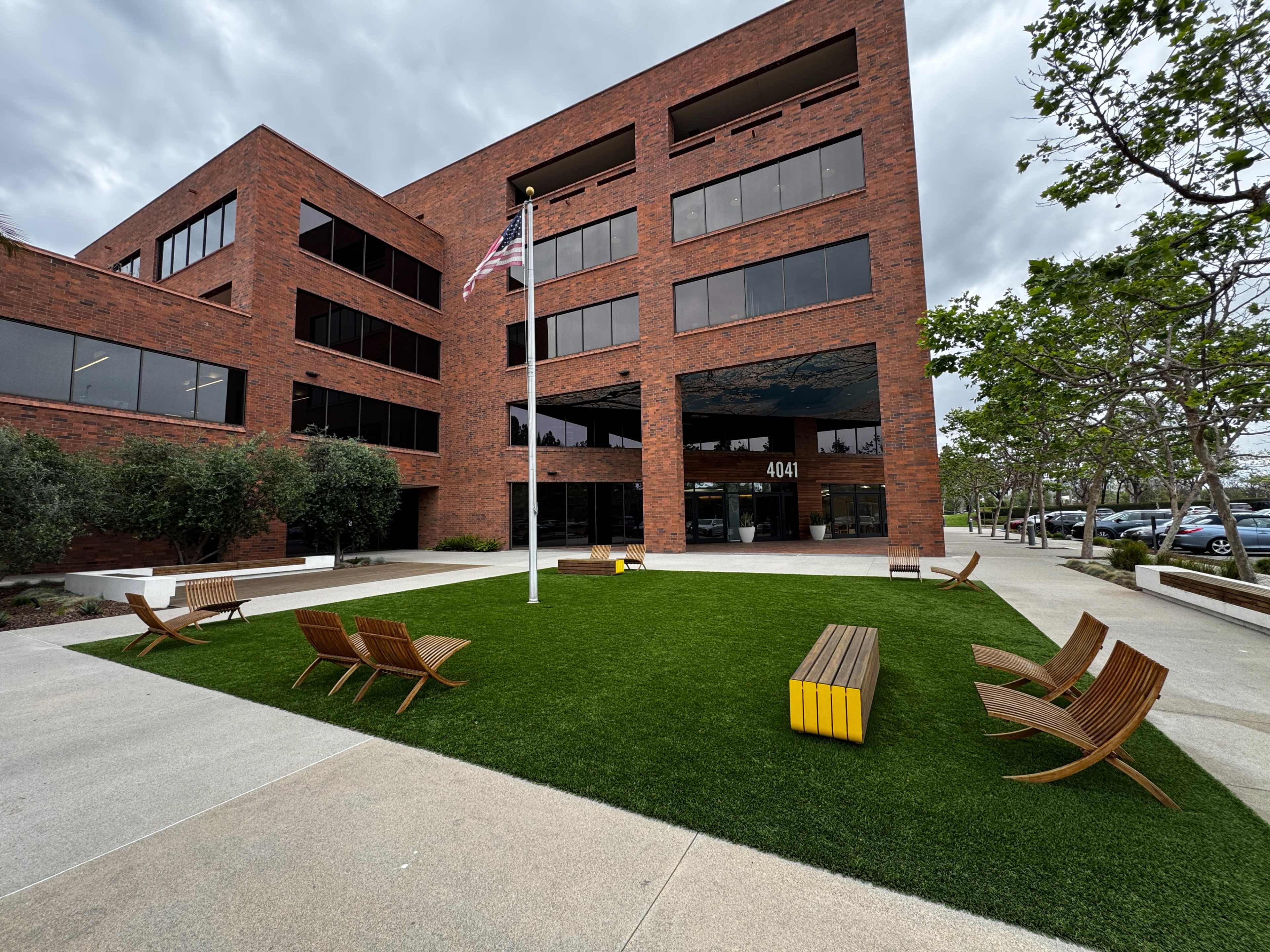 The image shows a modern brick office building with a grassy area featuring several wooden chairs and a flagpole.