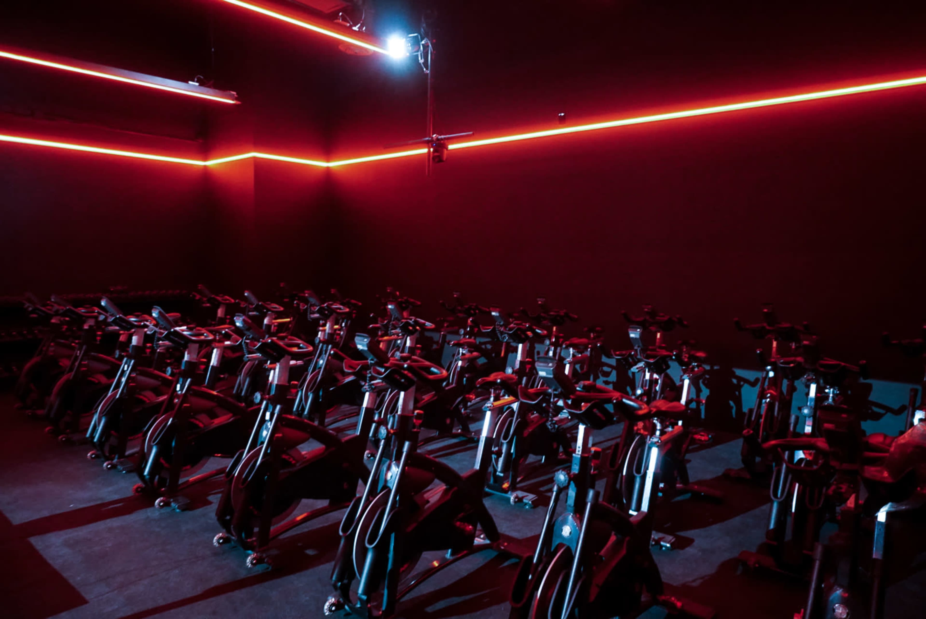 A dimly lit indoor cycling studio features rows of stationary bikes illuminated with red neon lights.