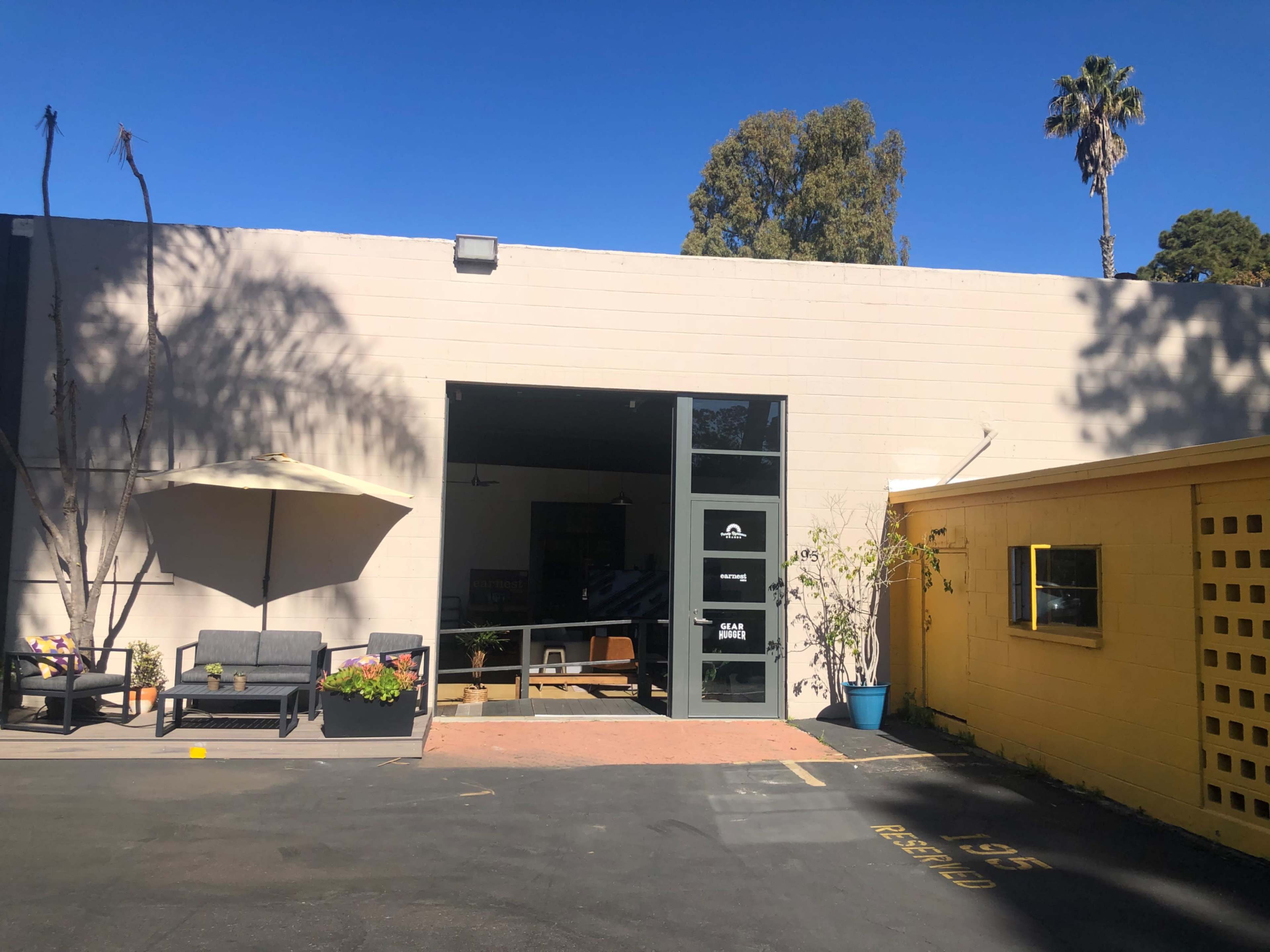 A gray building with large windows and an umbrella-covered seating area, alongside a smaller yellow building with a distinctive patterned exterior.