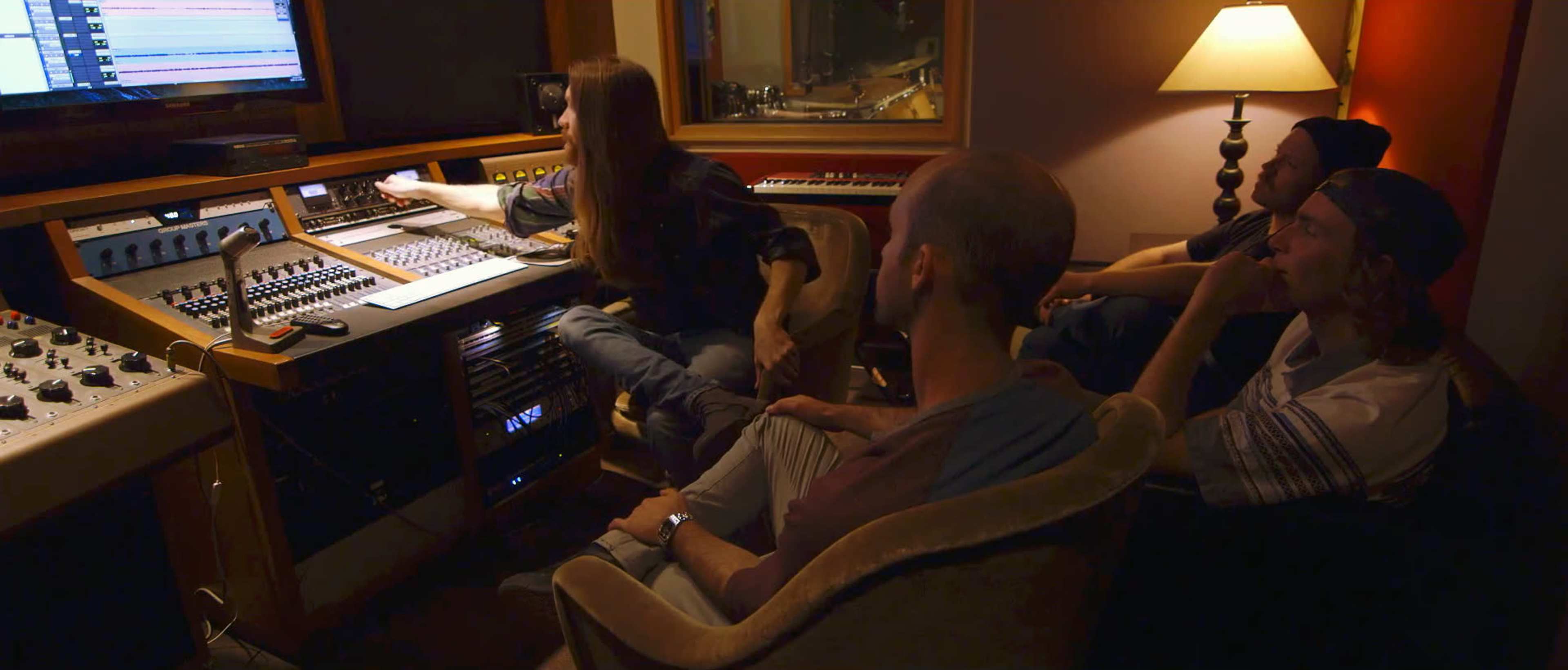A person adjusts audio equipment at a mixing console while three others sit on a couch observing in a recording studio.