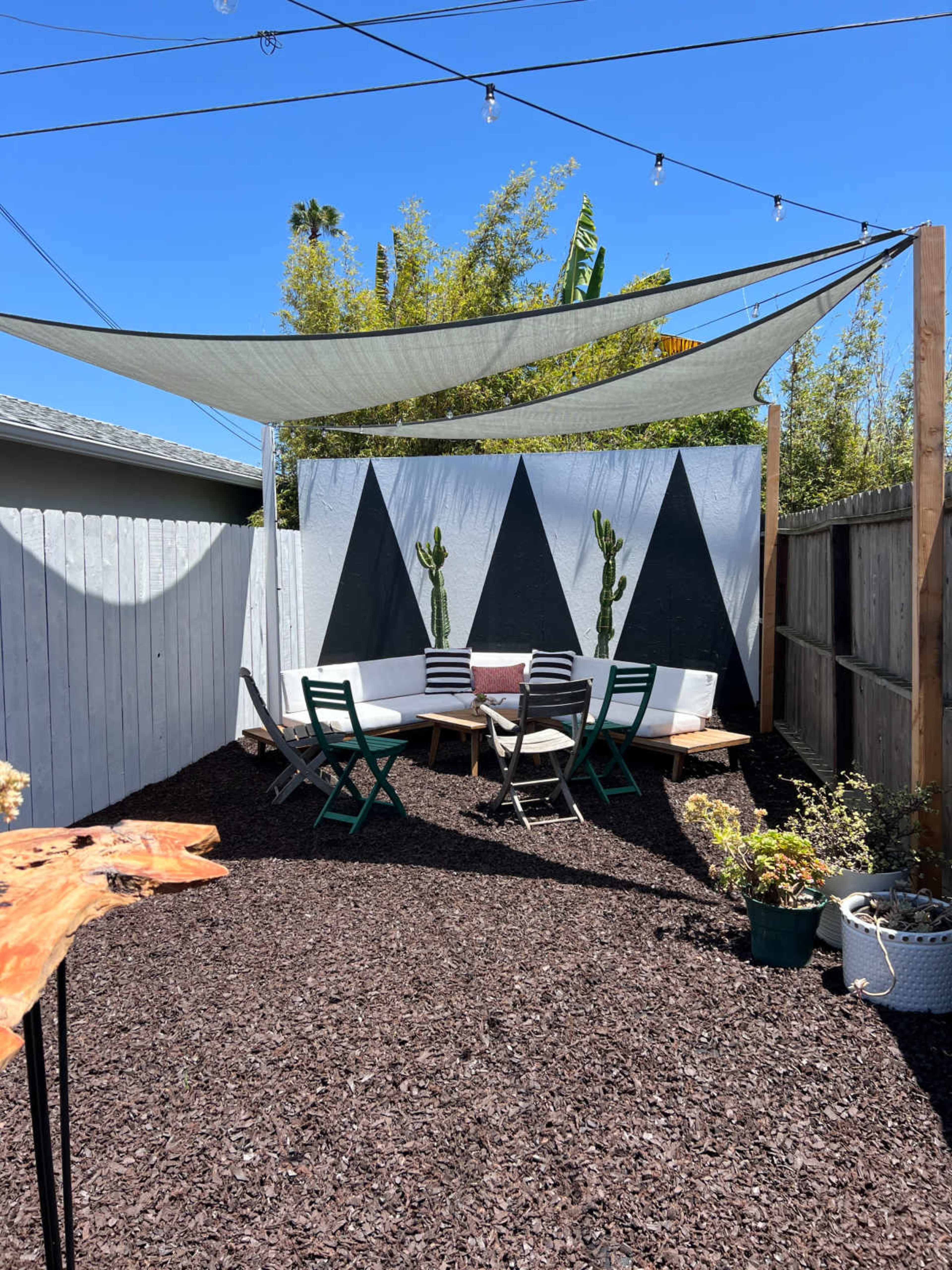 A shaded outdoor seating area with a geometric mural, surrounded by gravel and potted plants.