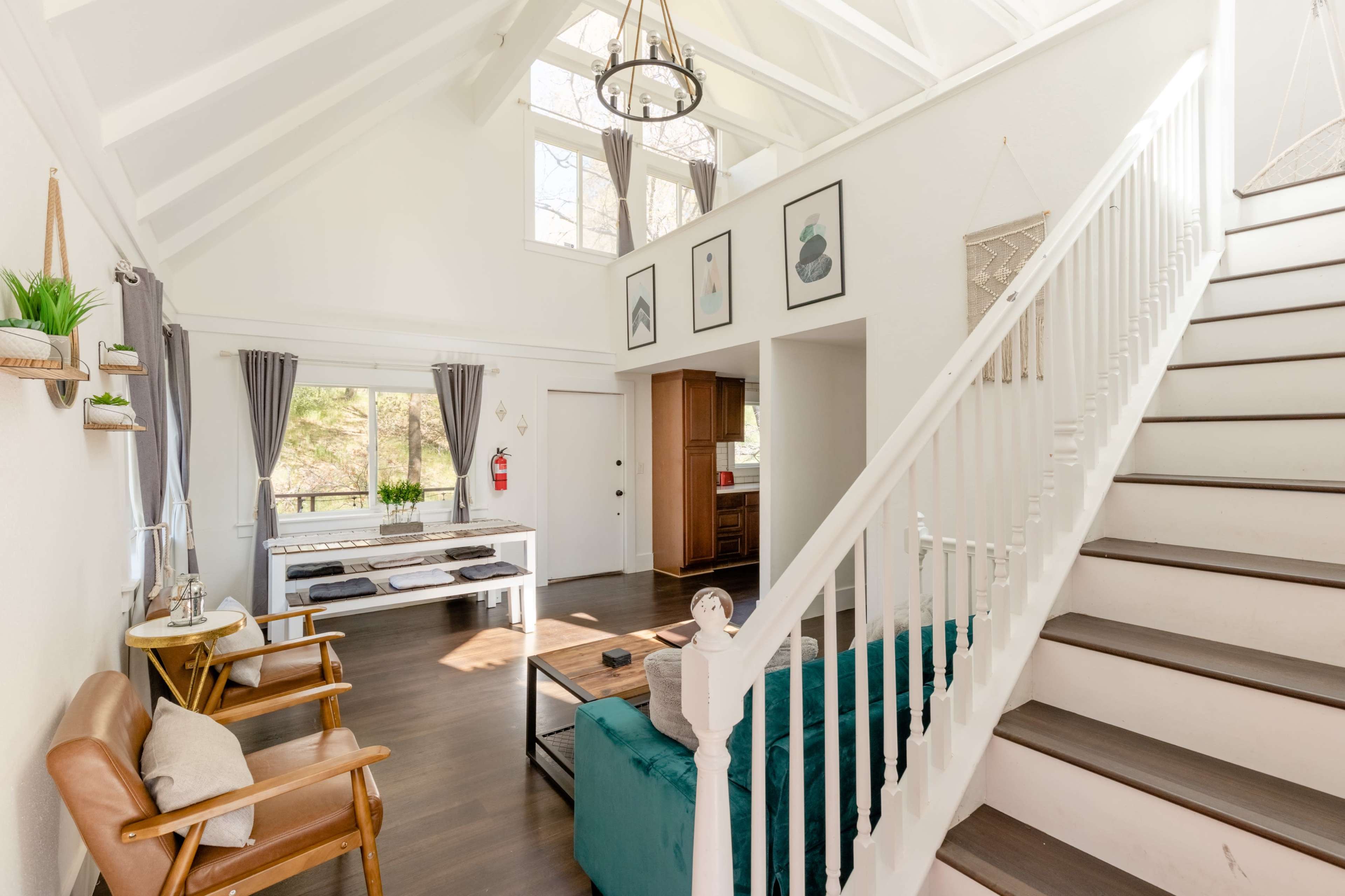 The image shows a bright, open living space with a high, sloped ceiling, featuring a stairway, a seating area with a teal sofa, and large windows letting in natural light.