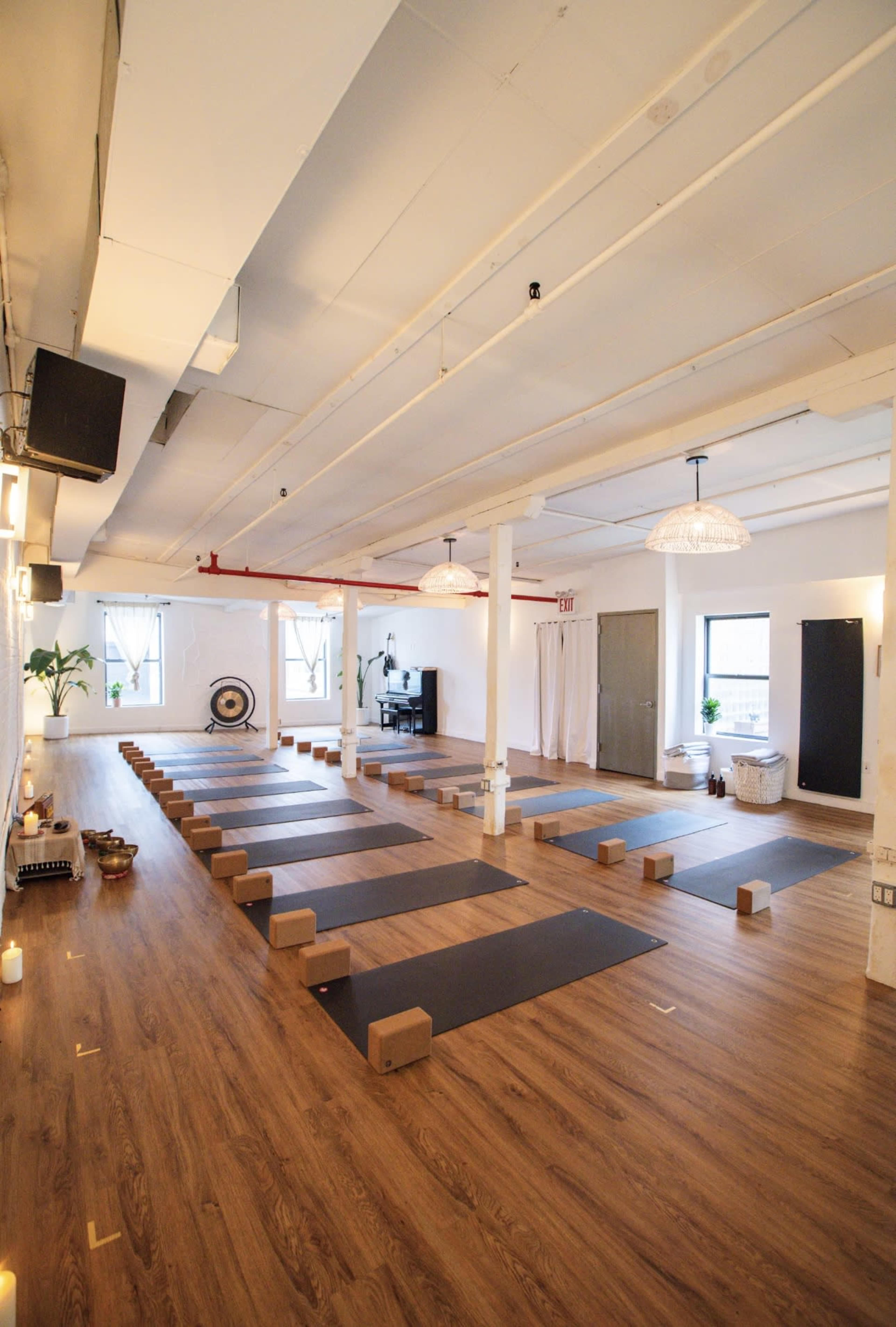 The image shows an empty yoga studio with mats aligned in rows, surrounded by wooden flooring and minimal decorations.
