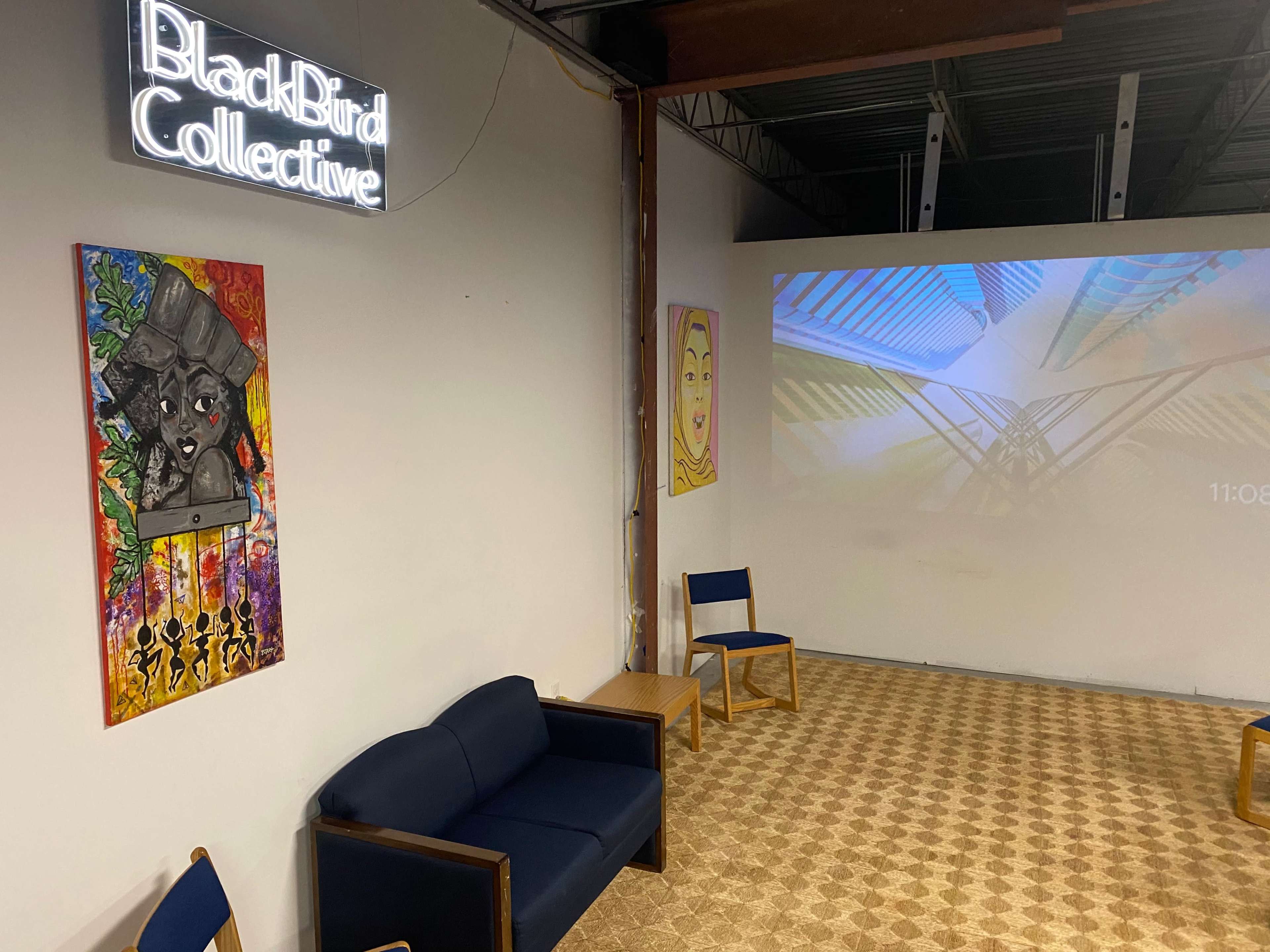 A room with two colorful wall paintings, a navy blue couch, wooden chairs, and a projection displaying abstract graphics on one wall.