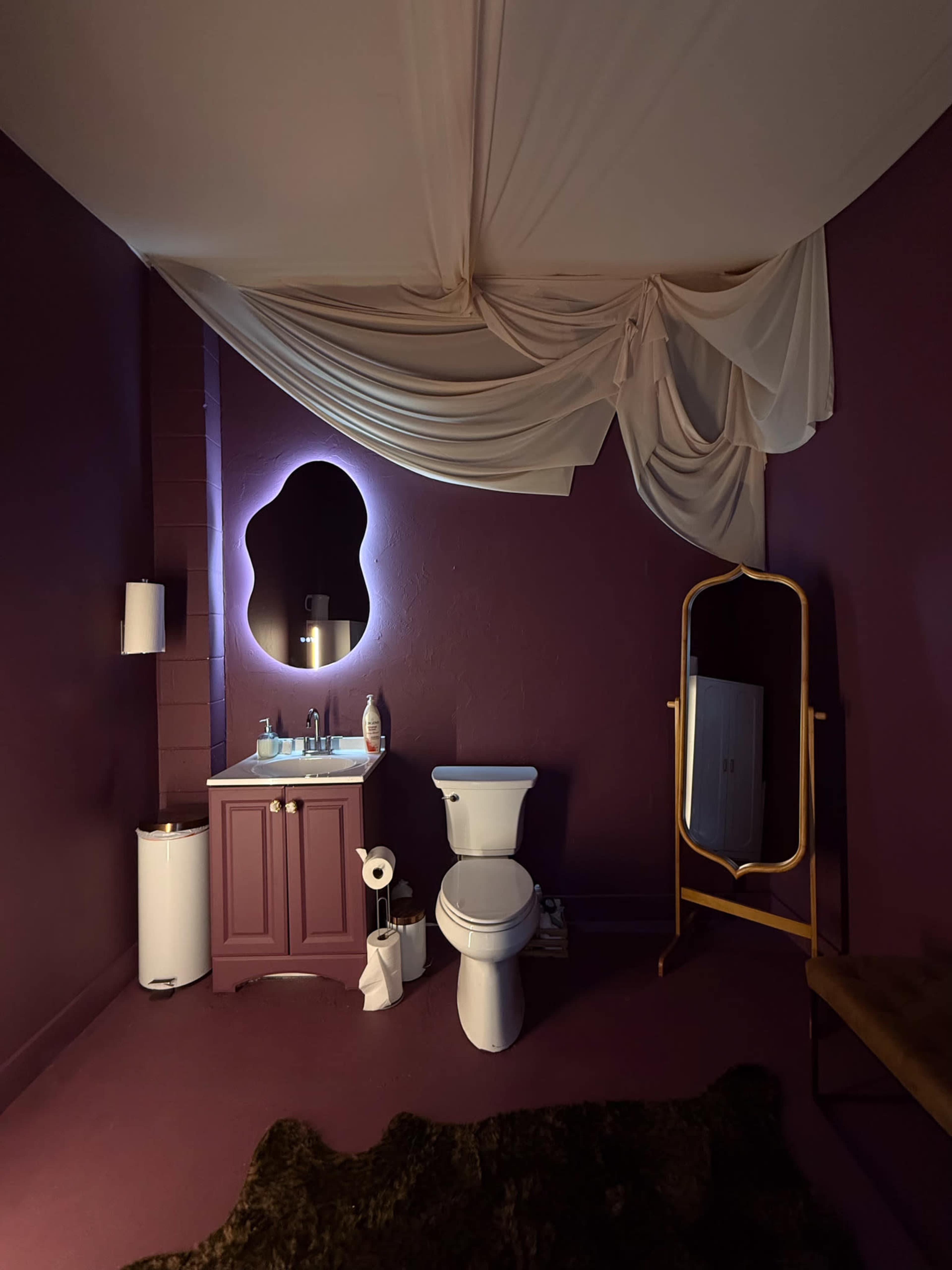 The image shows a small, purple bathroom with a vanity, a toilet, a full-length mirror, and a textured rug on the floor.