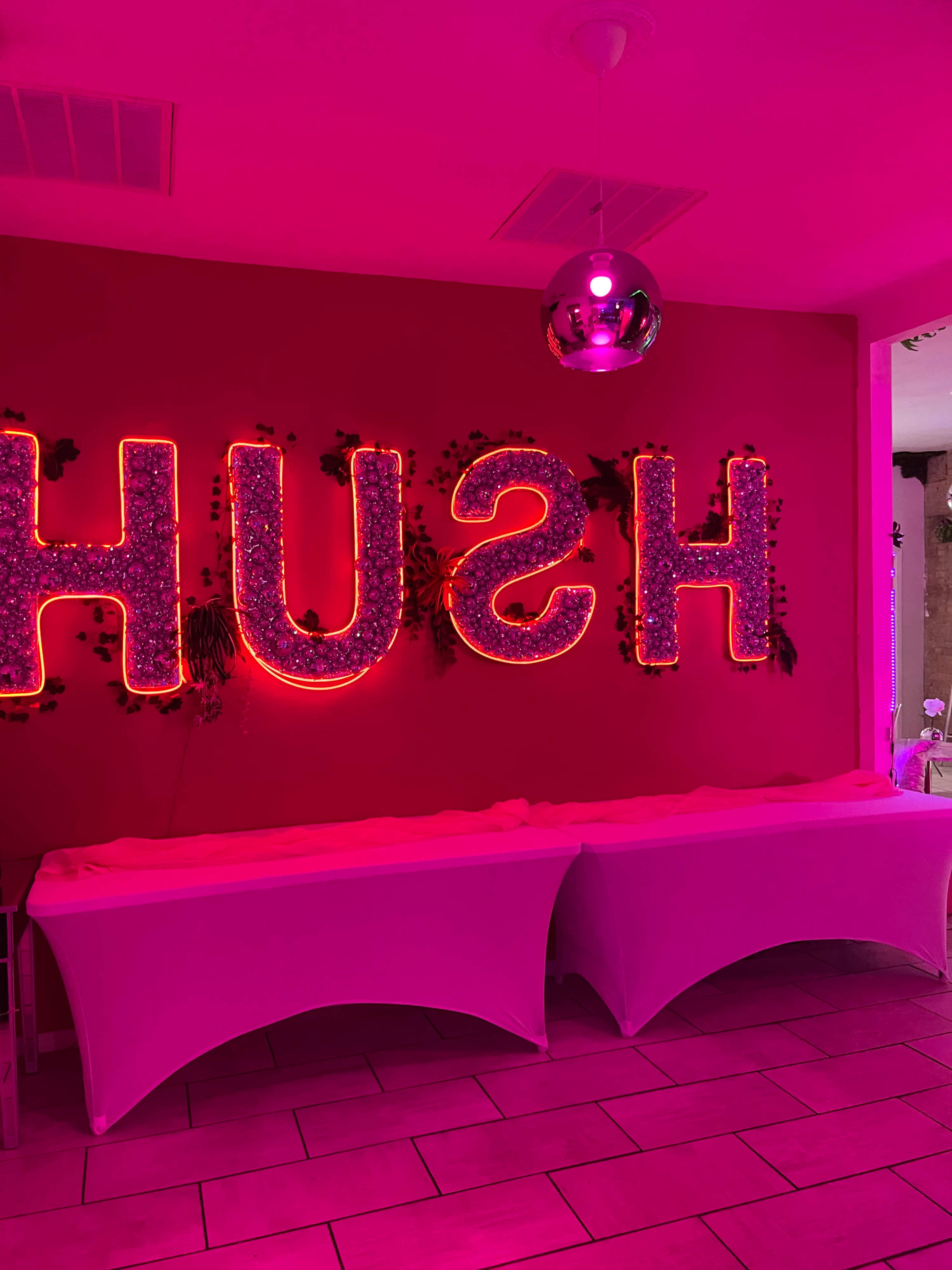 The interior features a wall with bright pink neon letters spelling "HUSH" against a contrasting red background, accompanied by a disco ball and white table coverings.