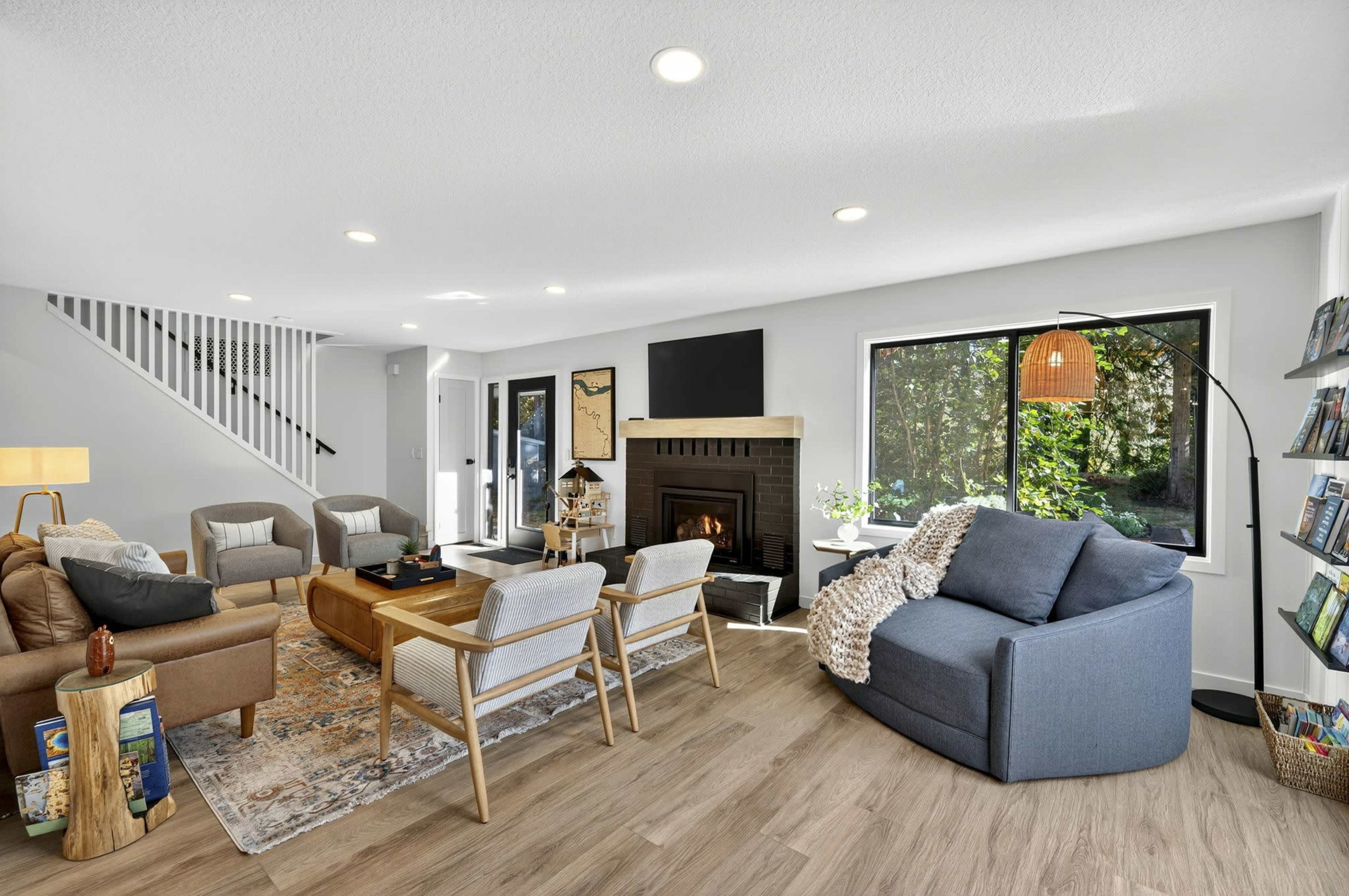 The image shows a modern living room with a fireplace, two sofas, a coffee table, and bookshelves, illuminated by natural light from large windows.