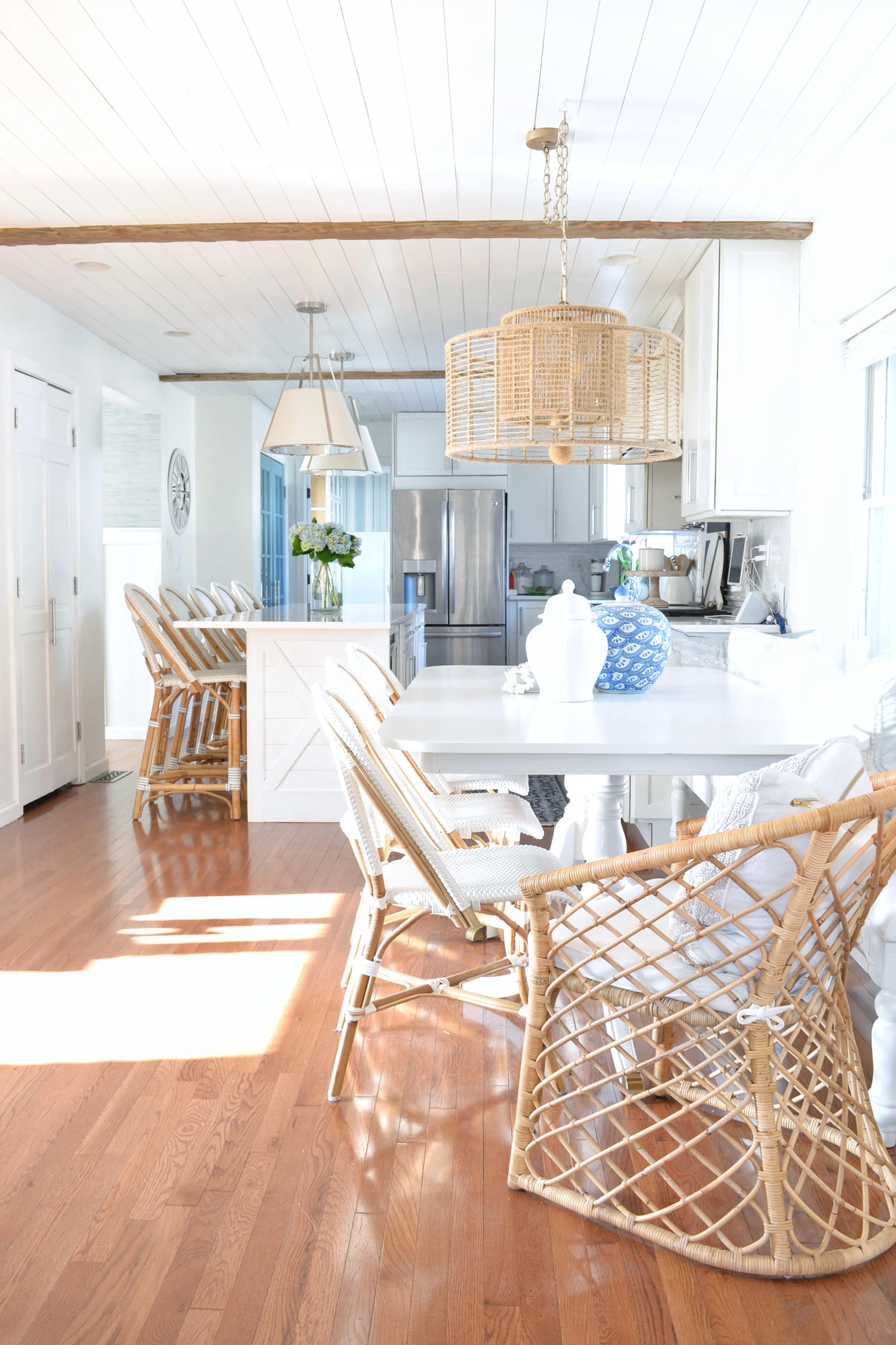 A bright, modern kitchen features a dining area with wooden flooring, a white table, and several wicker chairs arranged by the side.