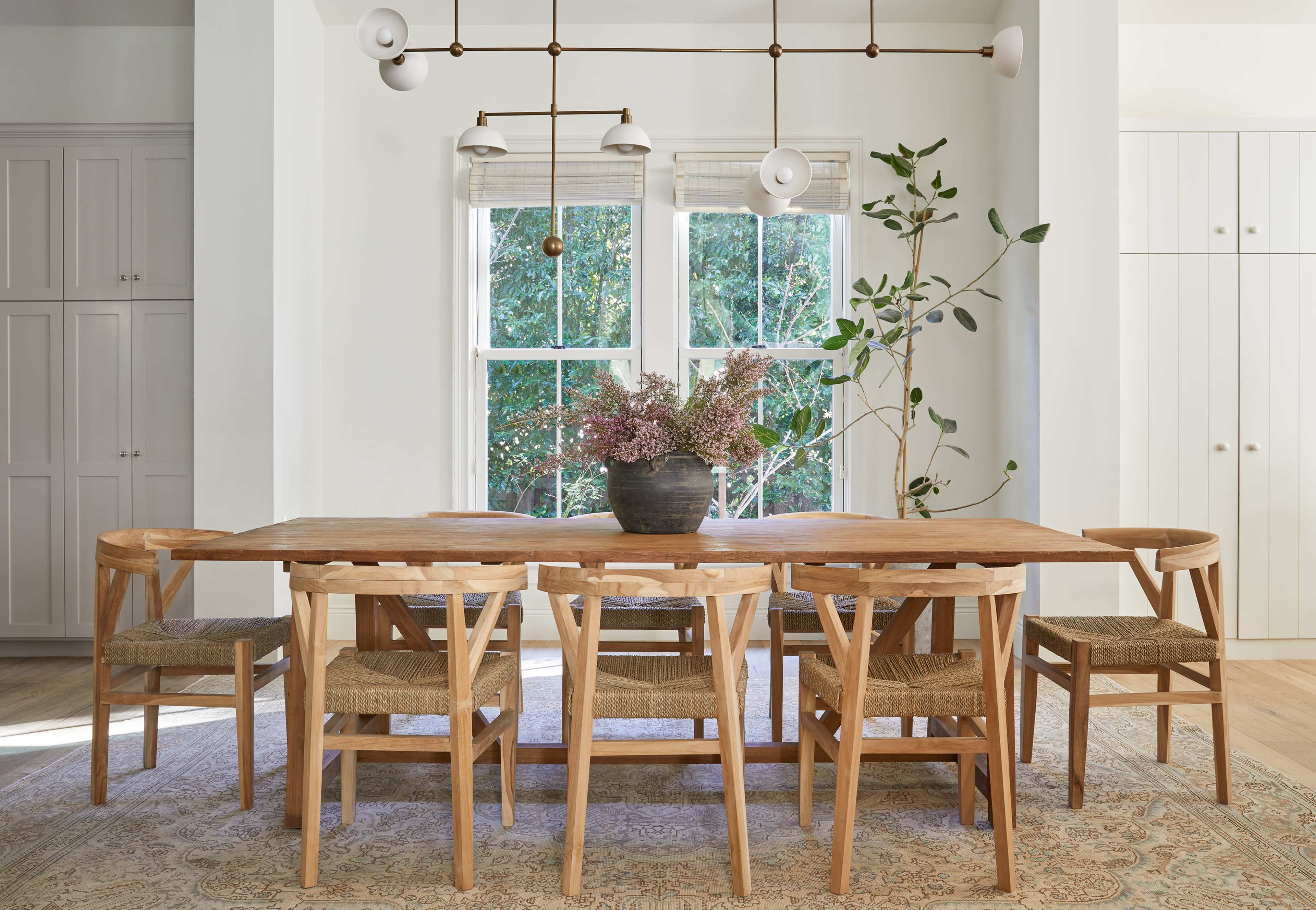 A wooden dining table surrounded by eight chairs is positioned in a bright room with large windows and a plant nearby.