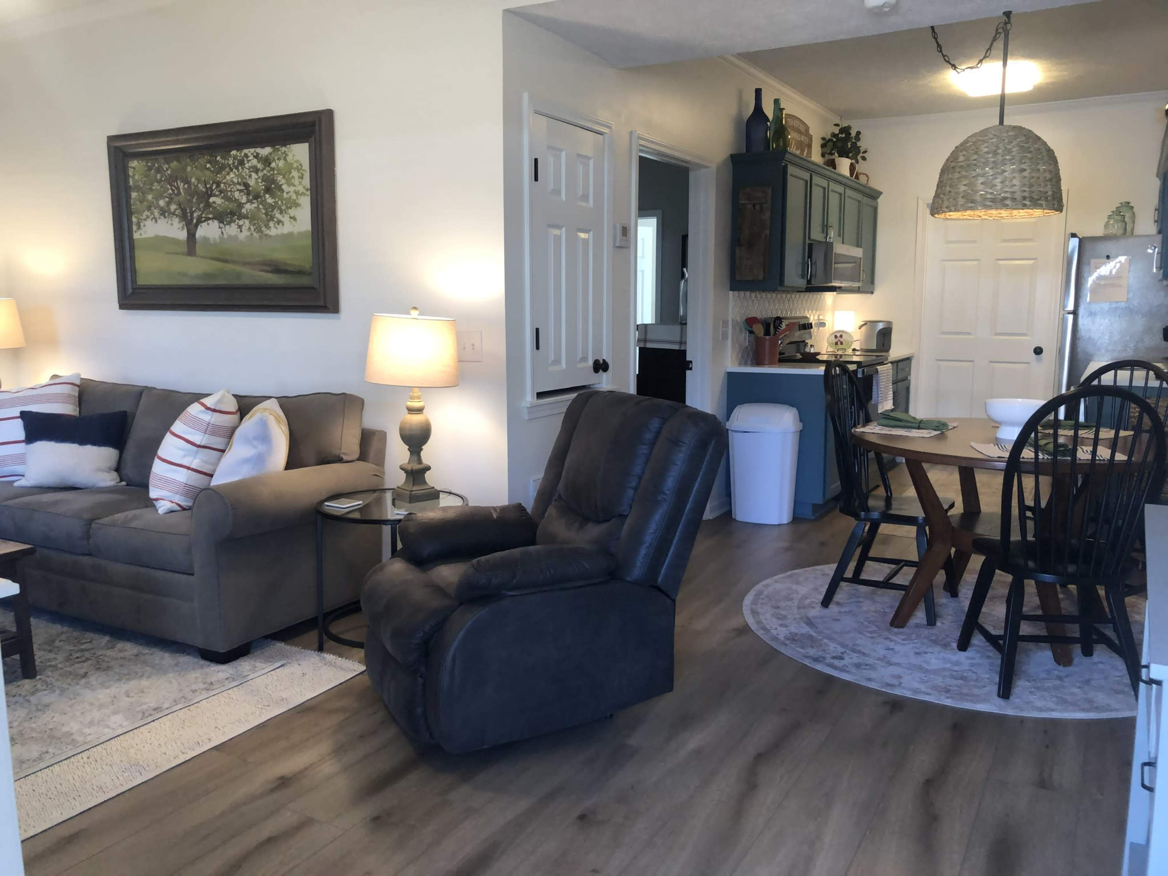 A living space featuring a gray sofa and a recliner, with a dining area that includes a round table and chairs, adjacent to a kitchen.
