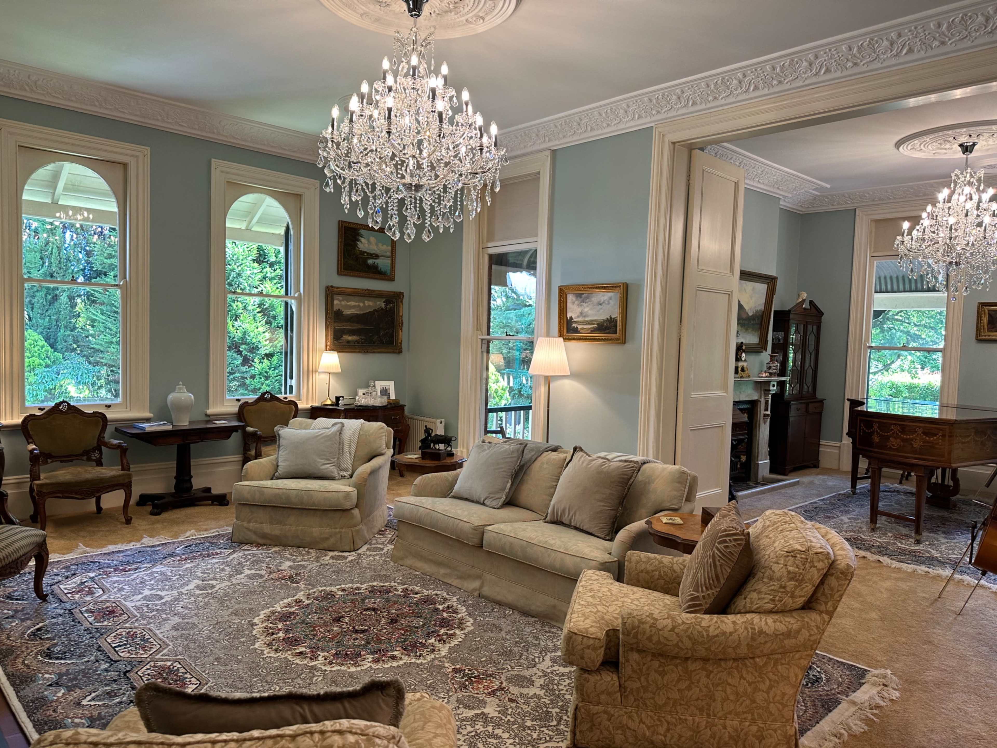 The room features two elegant chandeliers, a light blue color scheme, antique furniture, and large windows overlooking a garden.