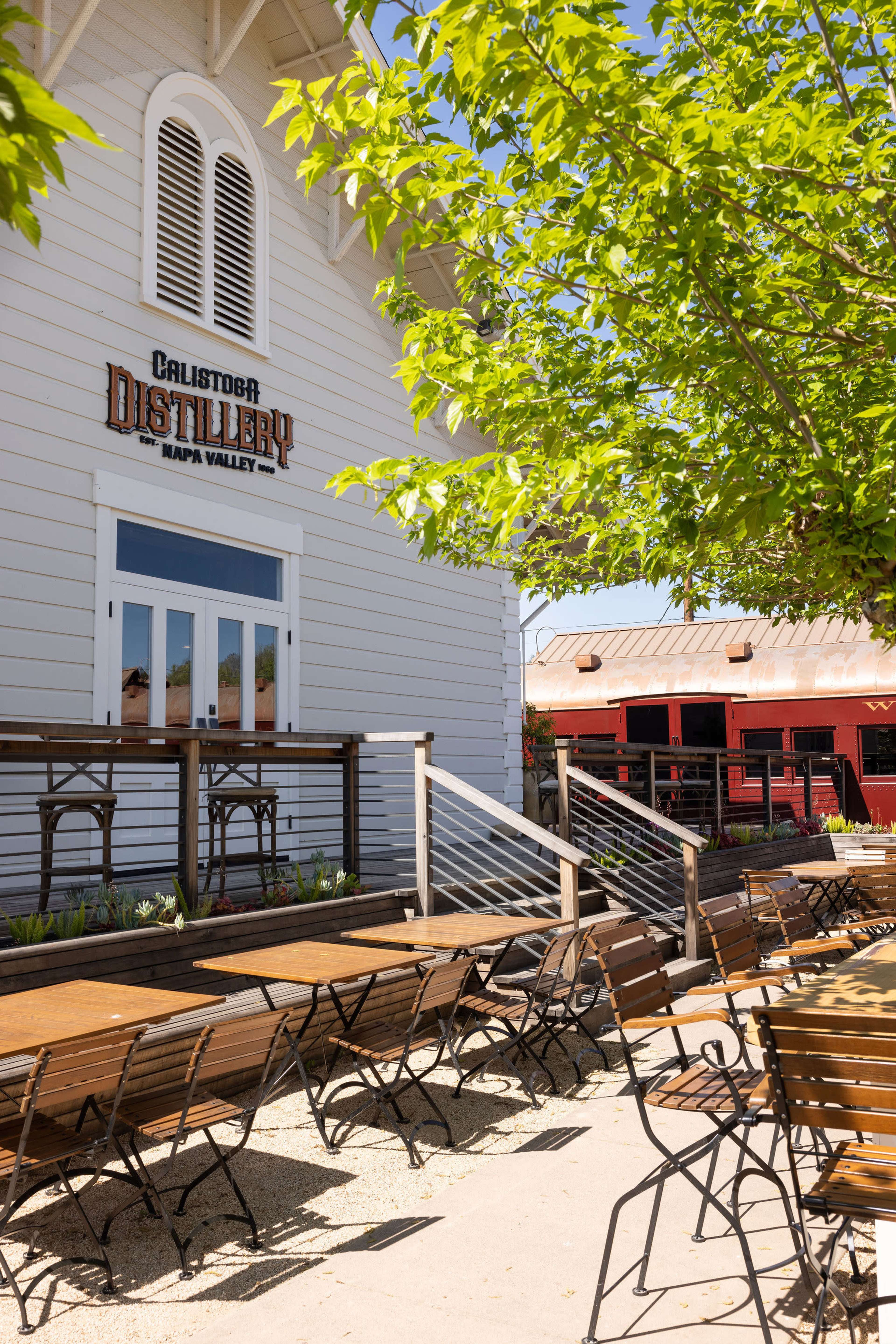 The image shows a white, wooden distillery building with a sign reading "Calistoga Distillery" and outdoor seating arrangements made of wooden tables and chairs under green trees.