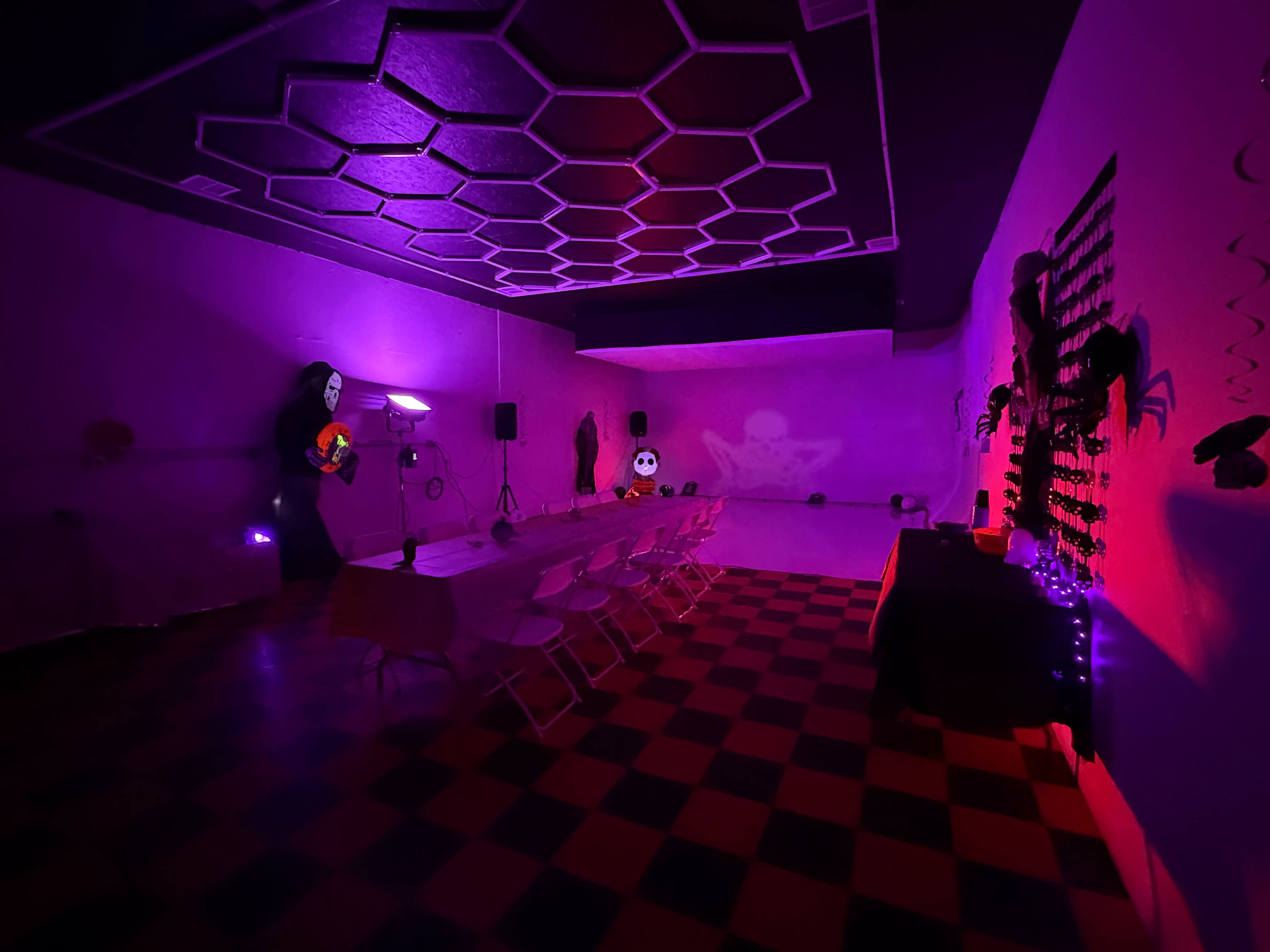 The image shows a dimly lit room decorated for a Halloween-themed event, featuring a checkered floor, a long table with chairs, and various spooky decorations along the walls.