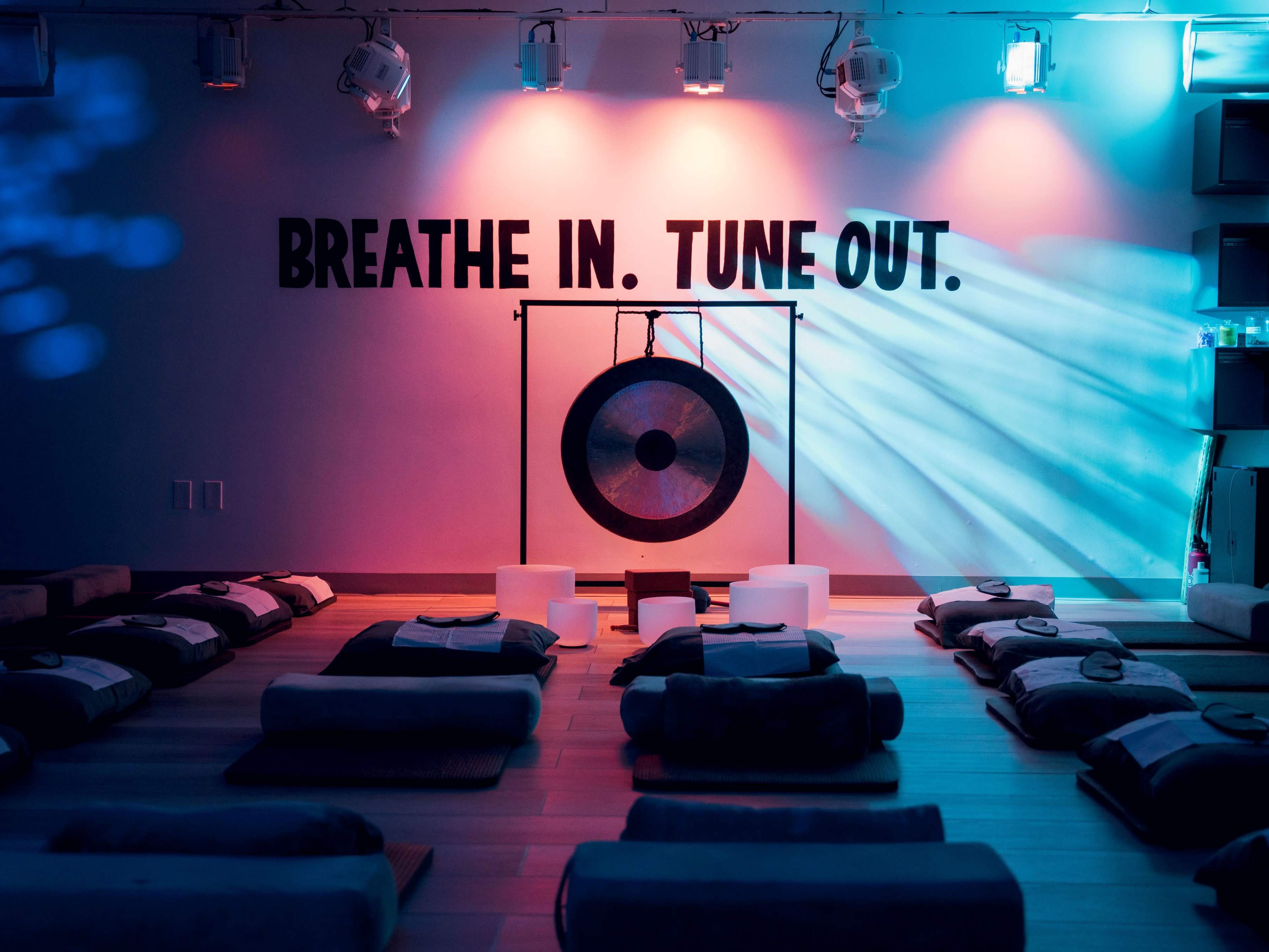 The image shows a meditation studio with floor cushions arranged in rows and a gong hanging on a stand, illuminated by colorful lighting and featuring the text "Breathe In. Tune Out" on the wall.