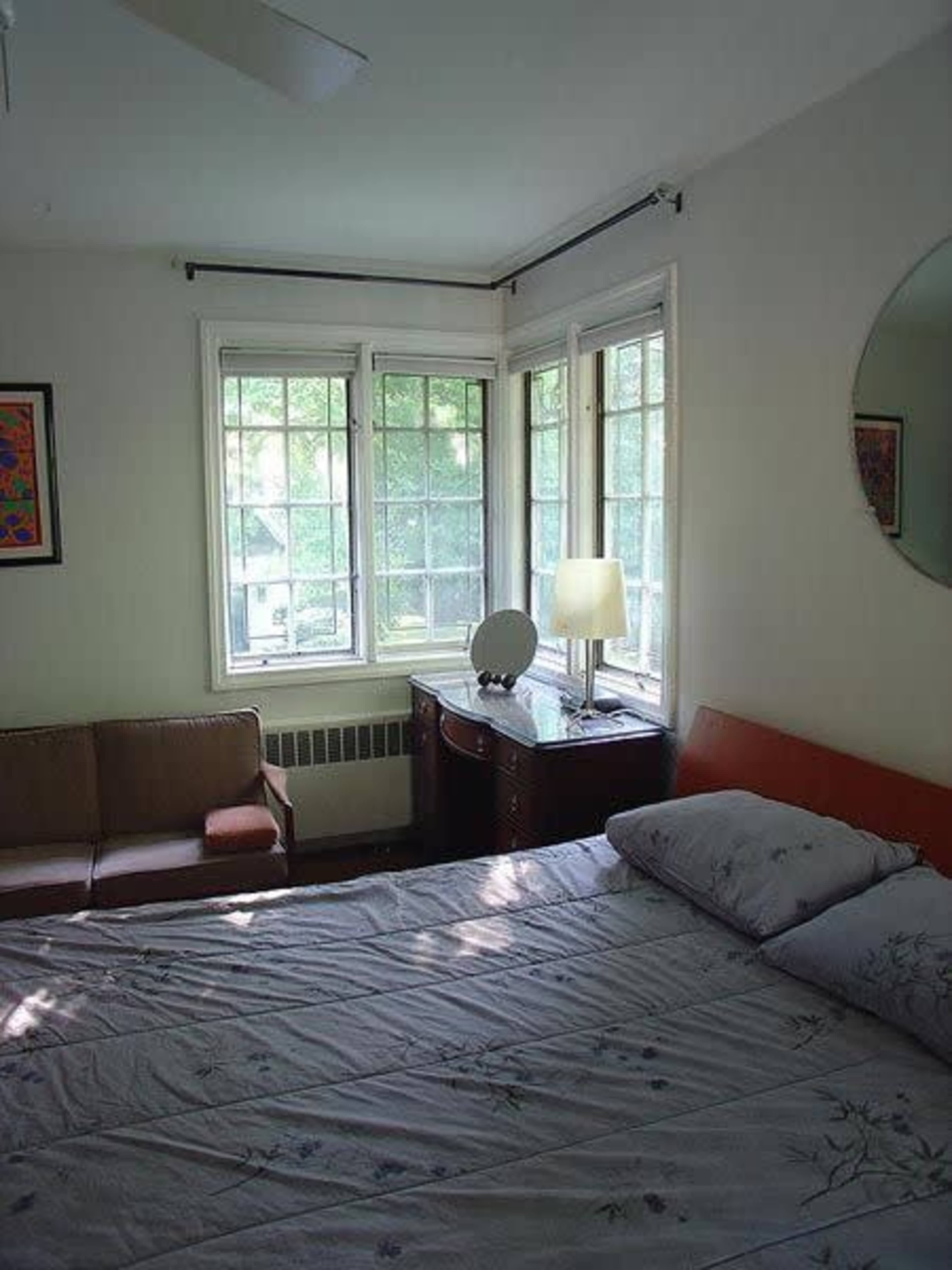 A small bedroom featuring a bed with floral-patterned sheets, a wooden nightstand with a lamp, and a seating area near large windows.