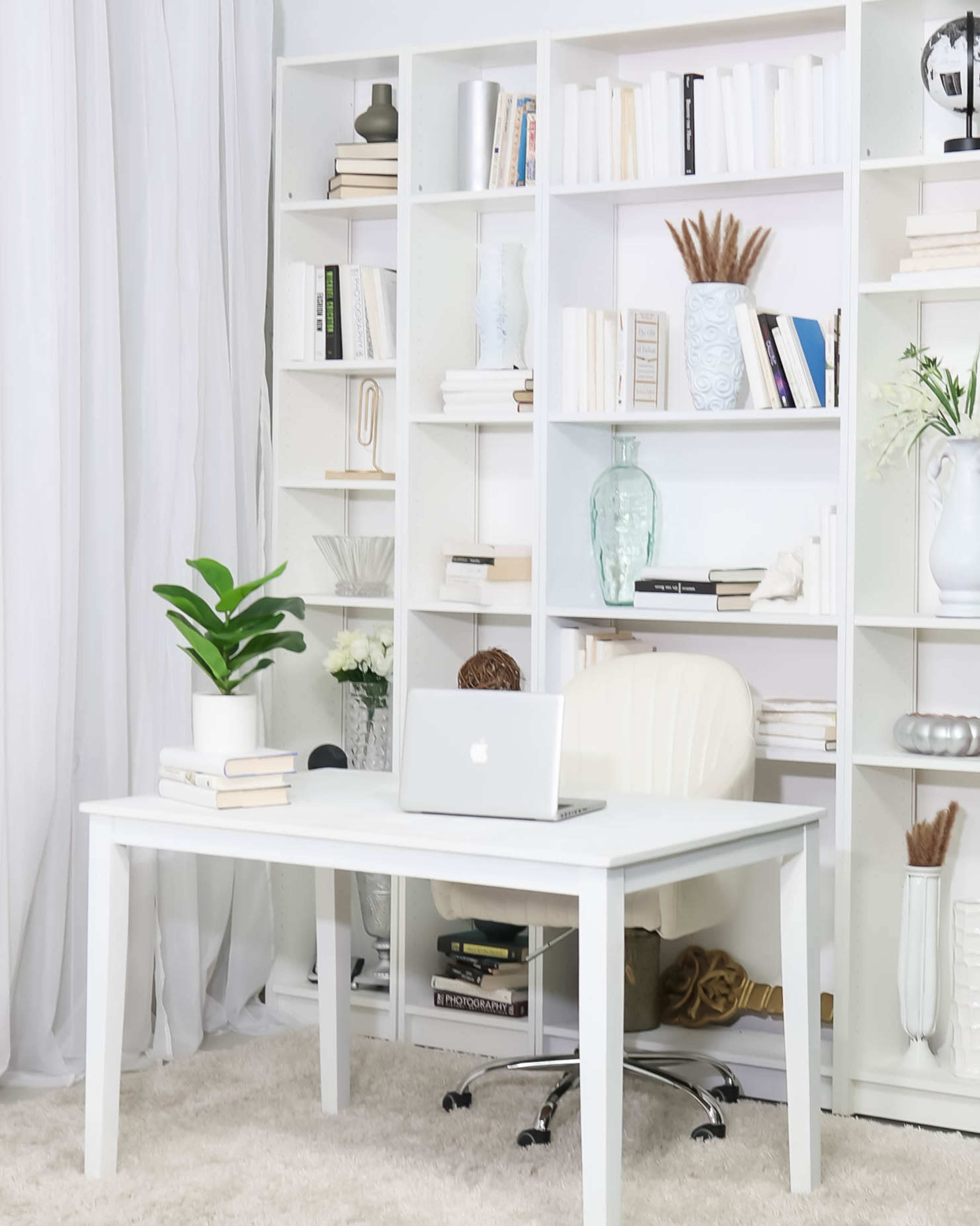 A minimalist home office features a white desk and chair in front of a bookshelf filled with books and decorative items, with a potted plant on the desk.