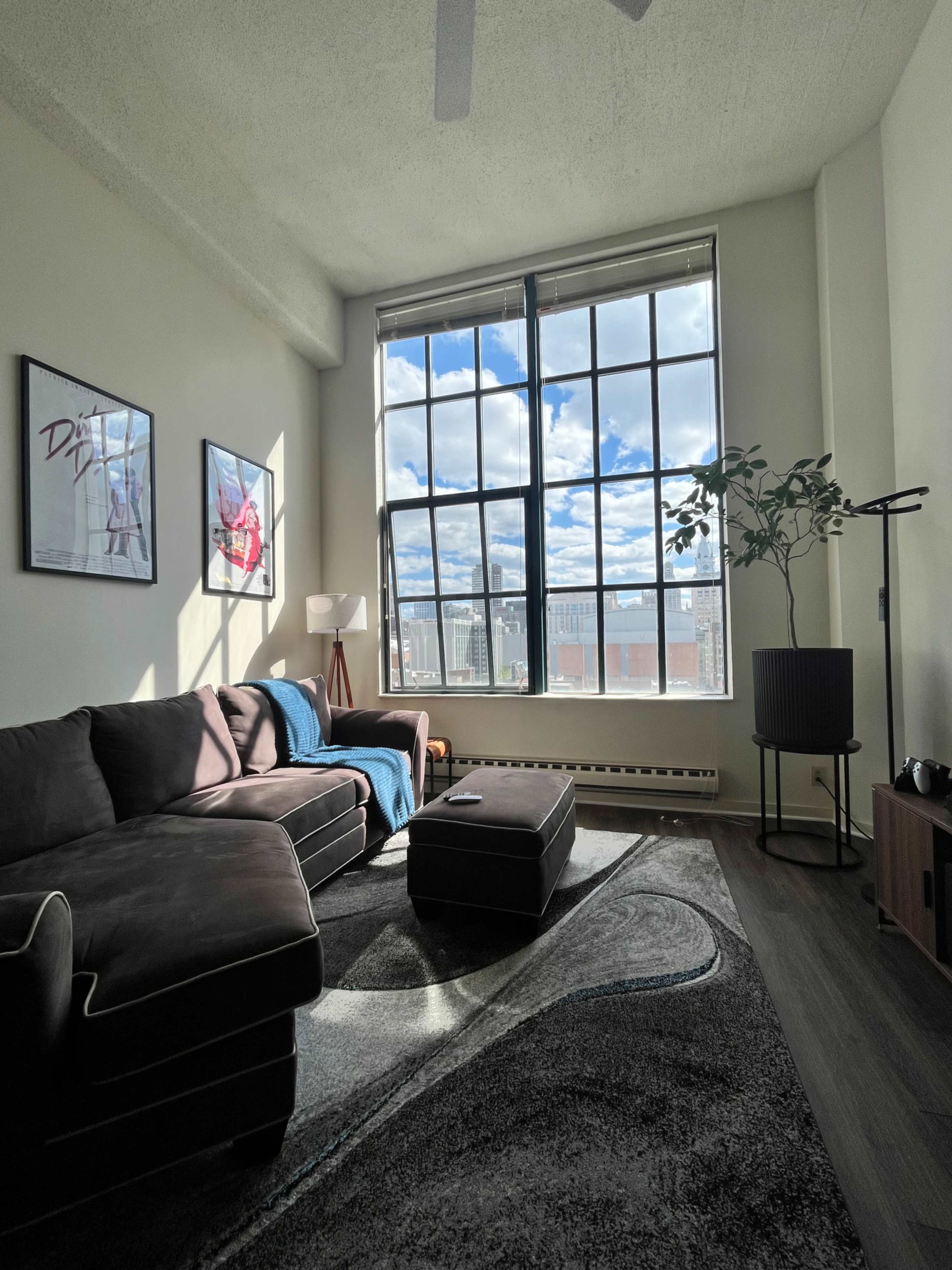 The image shows a well-lit living room with a large window, a dark sectional sofa, an ottoman, a lamp, and framed pictures on the wall.