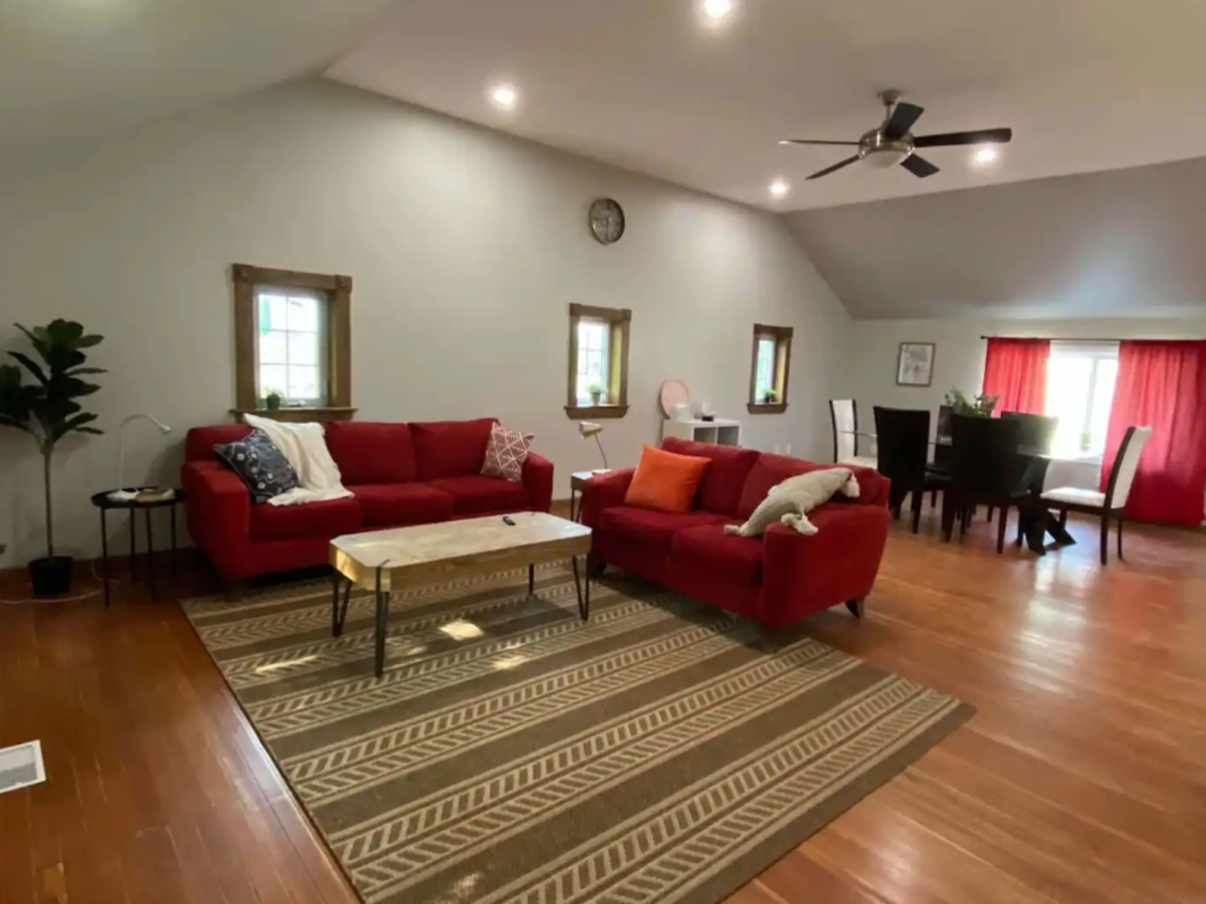 A spacious living room features a red sectional sofa, a marble coffee table, and a dining area with a wooden table, all illuminated by overhead lighting.