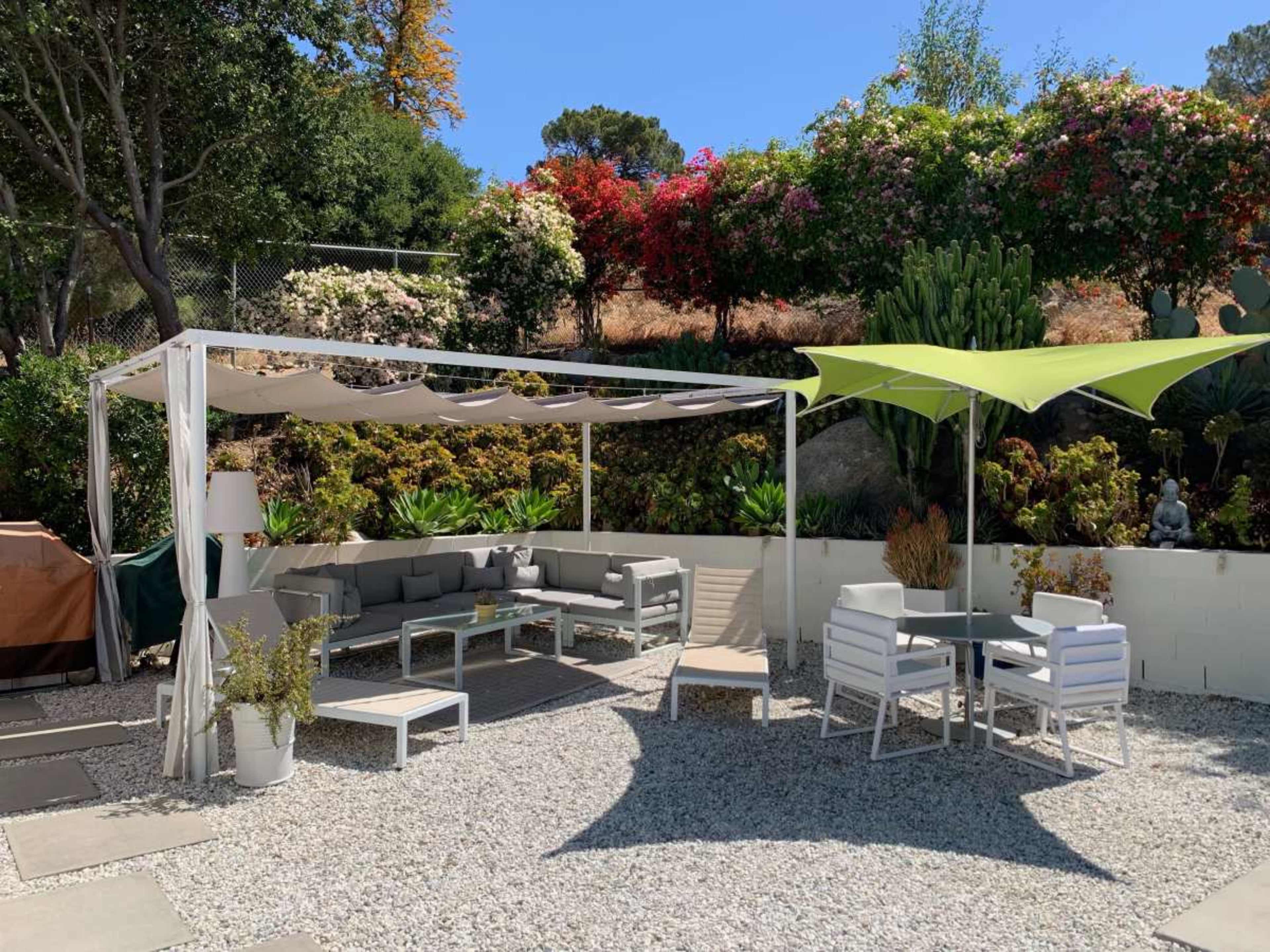 The image shows a landscaped outdoor patio area featuring a shaded seating arrangement under a canopy, surrounded by various plants and colorful flowers.