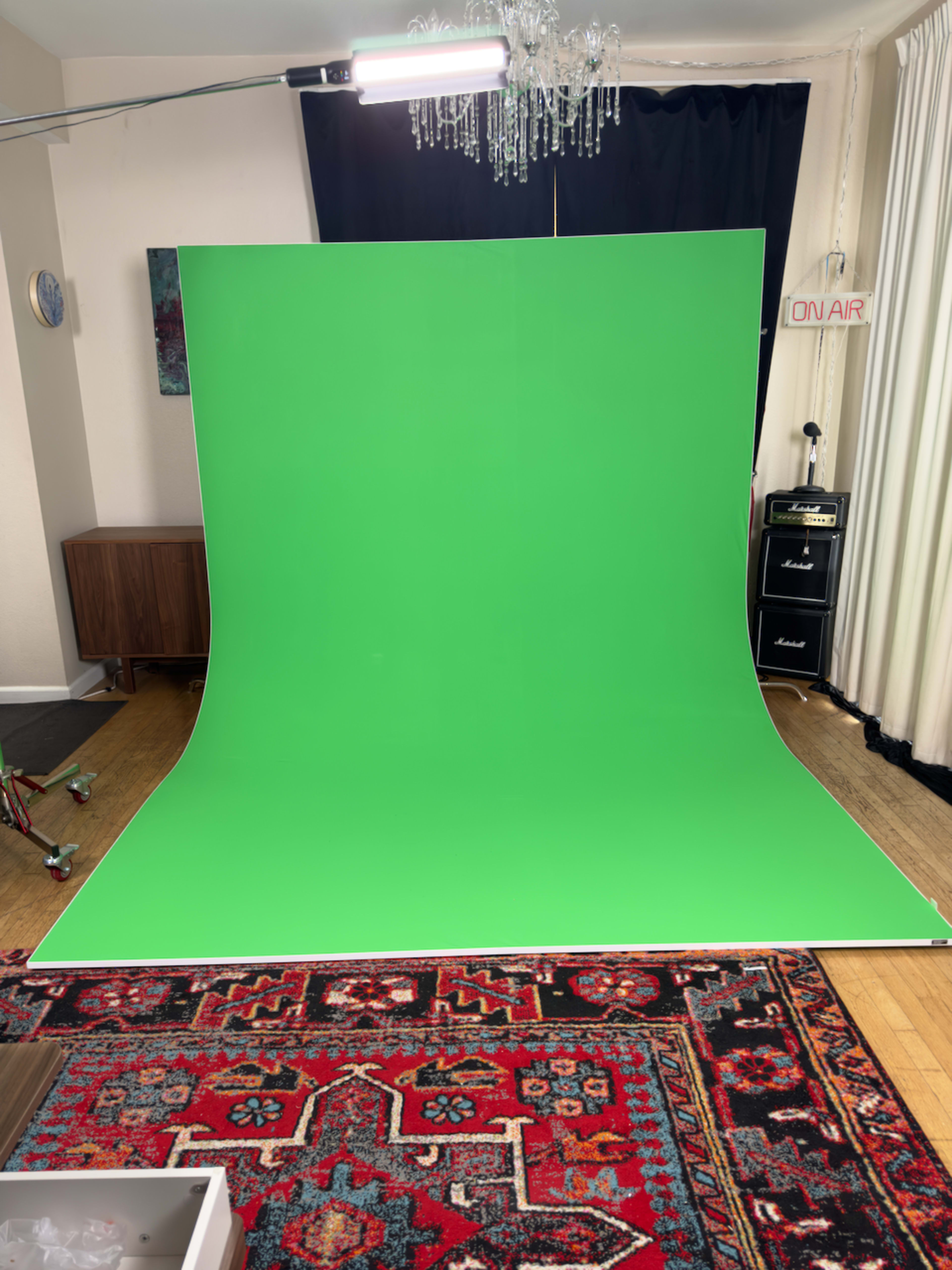 The image shows a green screen setup in a room, featuring a large green backdrop on the floor, a decorative rug, and equipment like a light and a speaker cabinet.