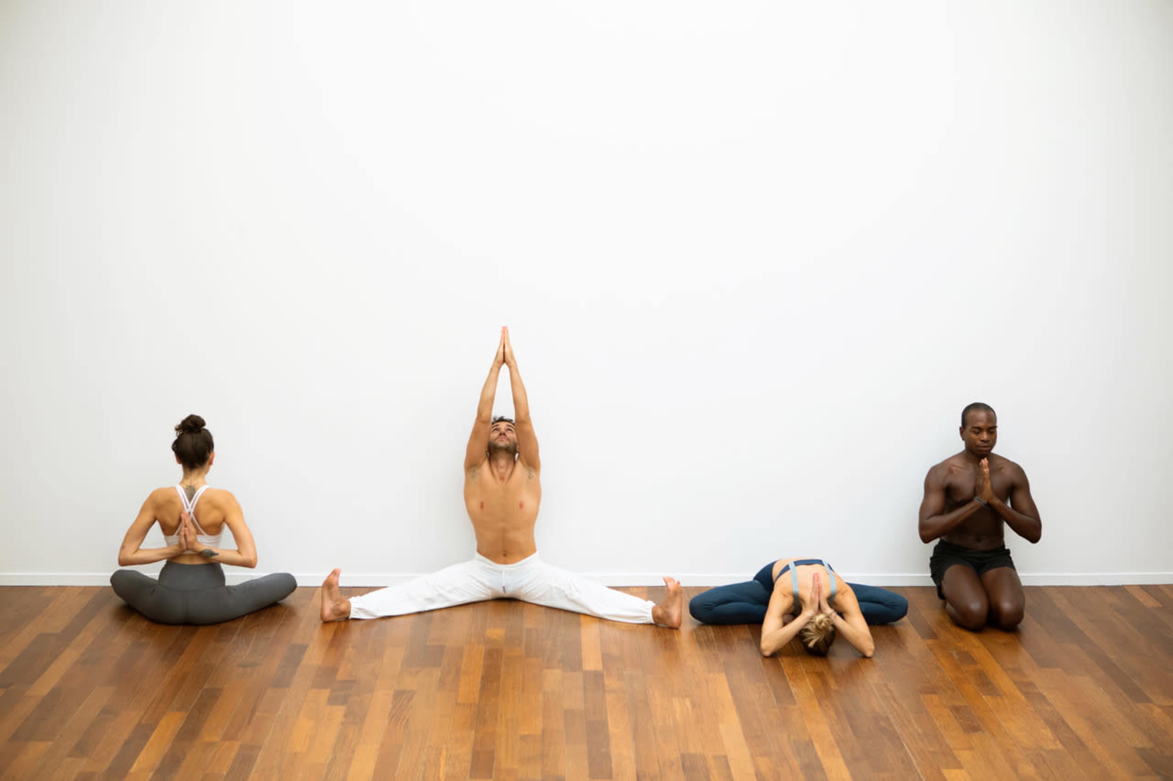 Four individuals practice yoga on a hardwood floor, each in a different pose against a plain white wall.