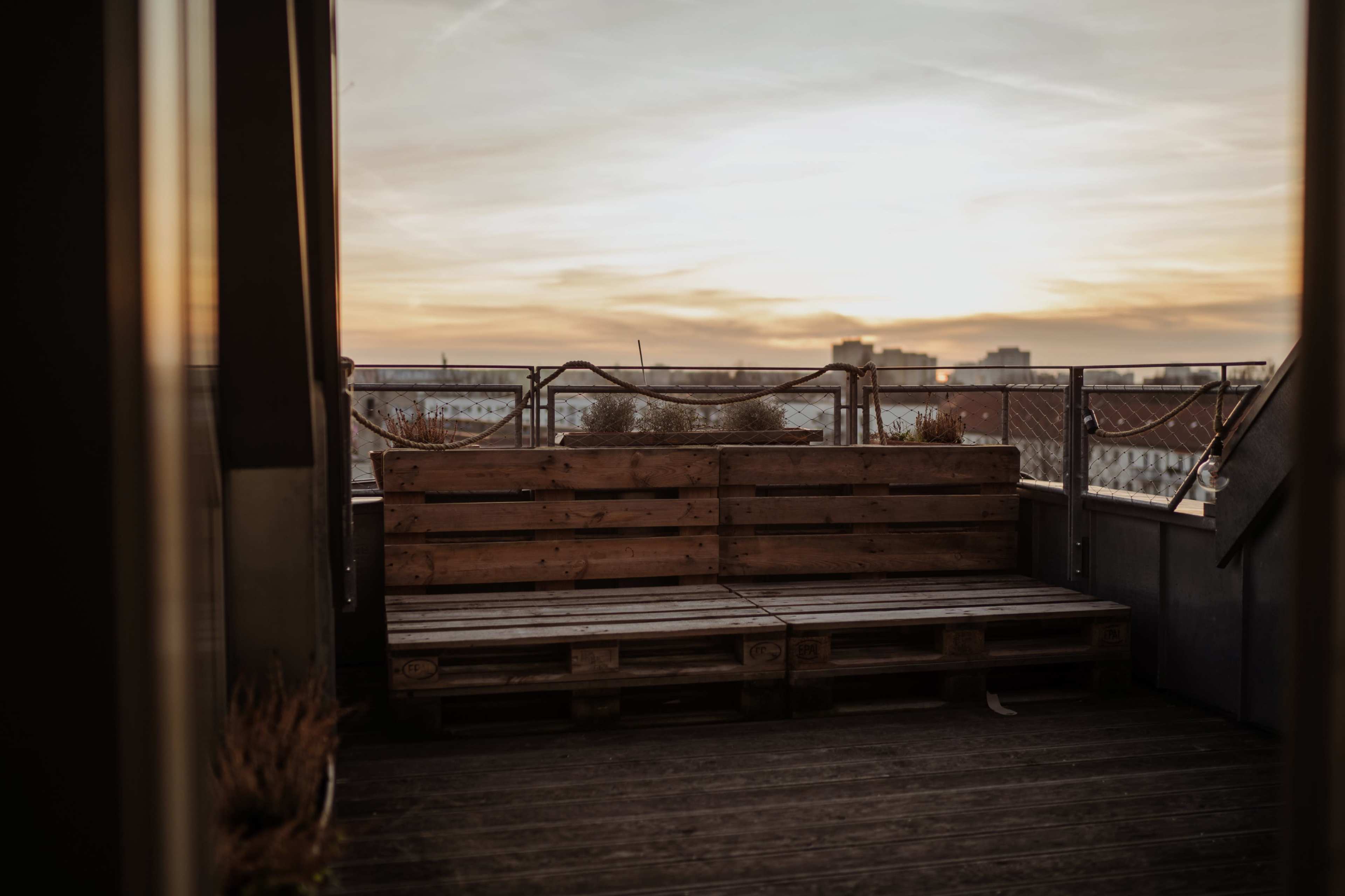 A wooden pallet bench is positioned on a balcony overlooking a sunset-lit sky with distant buildings.