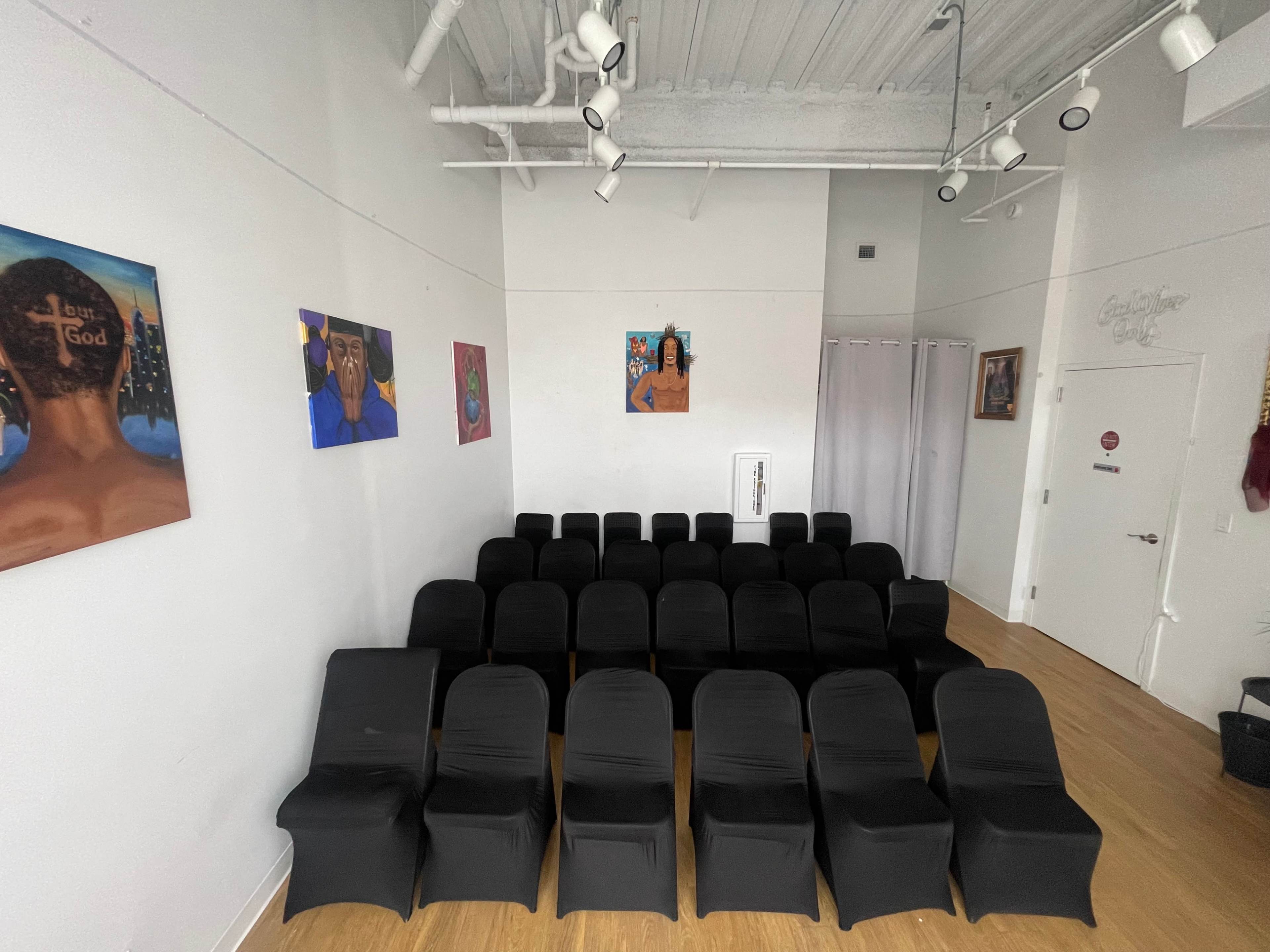 The image shows a row of black chairs arranged neatly in a spacious, well-lit room featuring colorful artwork on the walls.