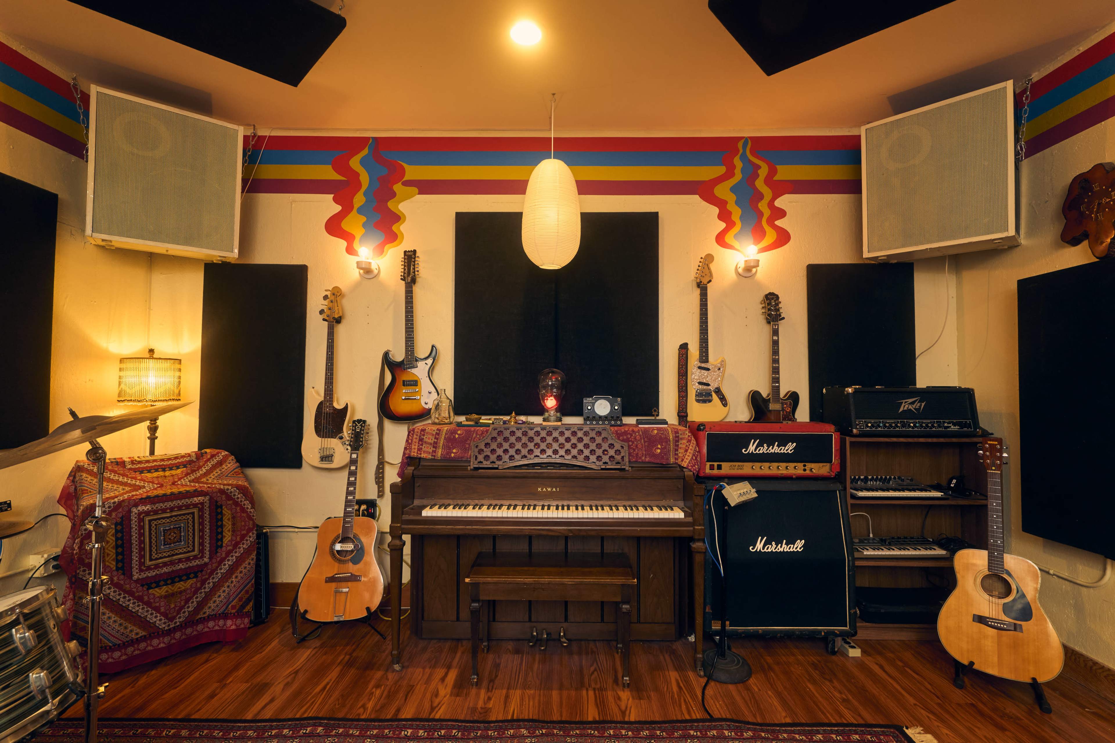 The image depicts a well-equipped music studio featuring guitars, amplifiers, a keyboard, and decorative wall elements.