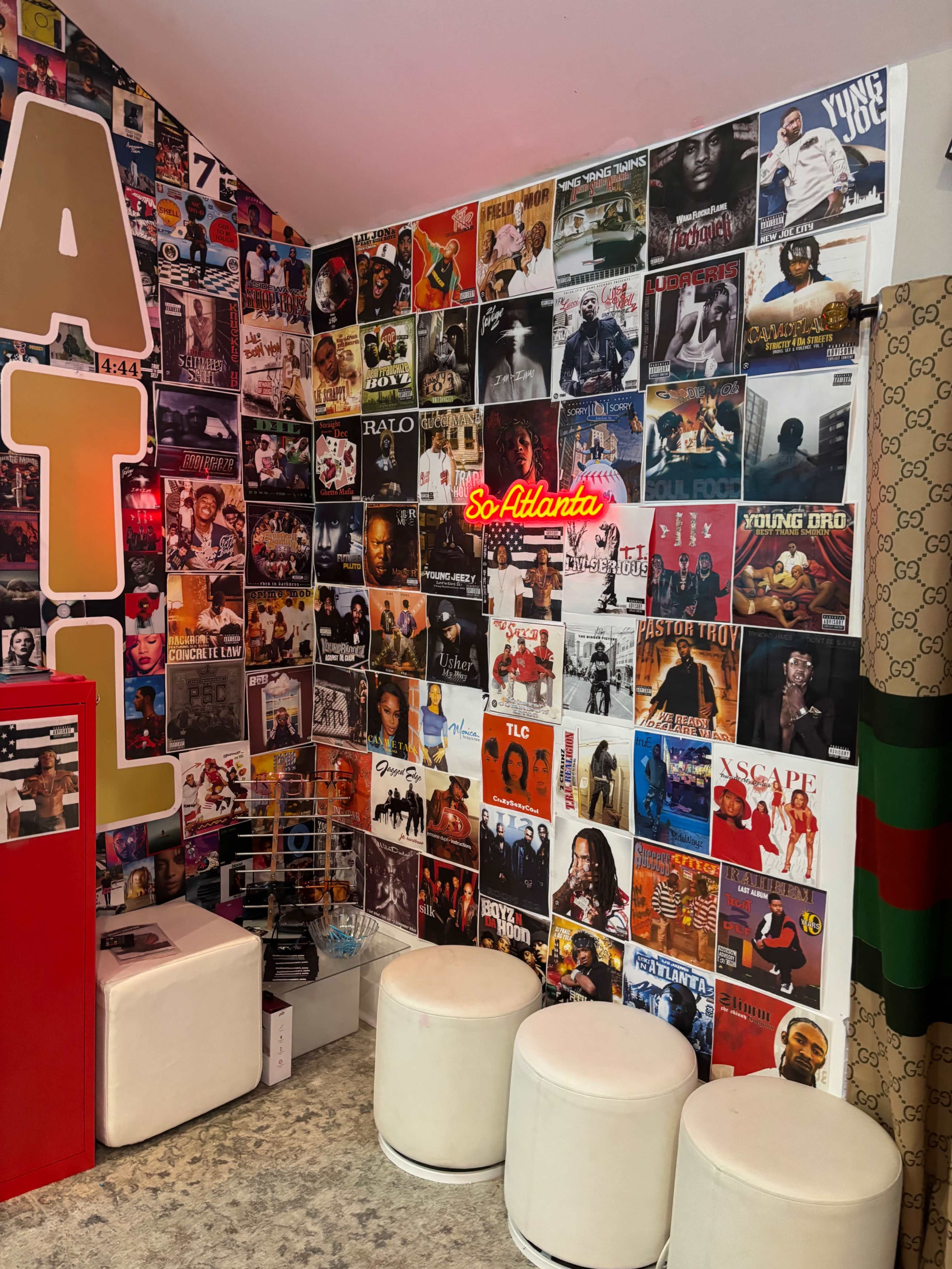 The image shows a corner of a room adorned with a collage of music posters and a neon sign that reads "So Atlanta," alongside two white stools.