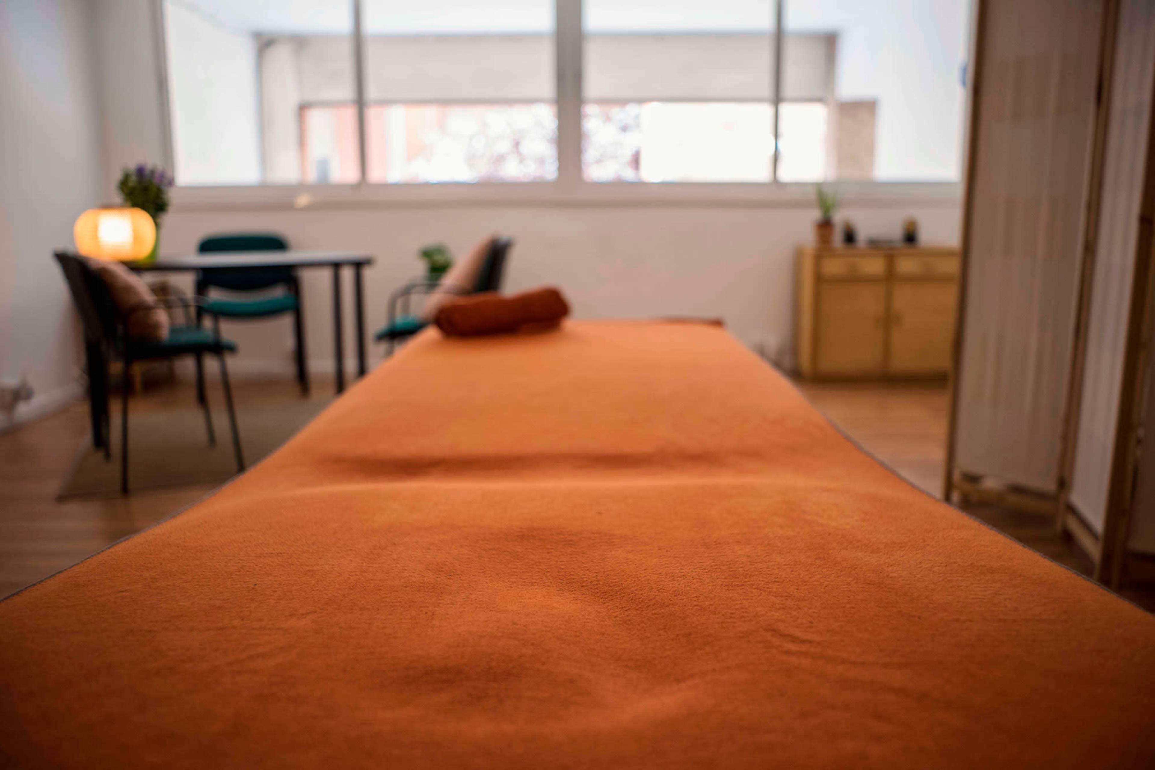 A cozy treatment room with an orange massage table, a small table with chairs, and a wooden cabinet in the background.