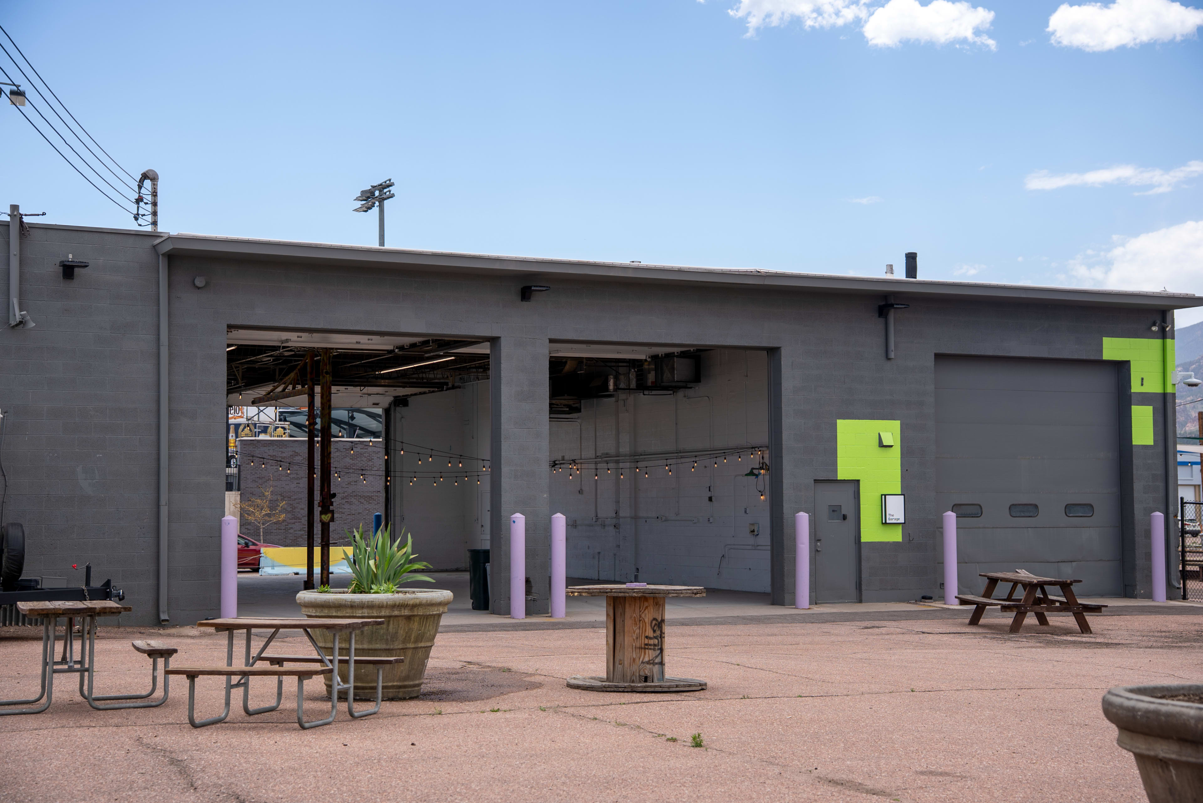 The image shows a gray building with two open garage doors, surrounded by outdoor seating and overhead string lights.