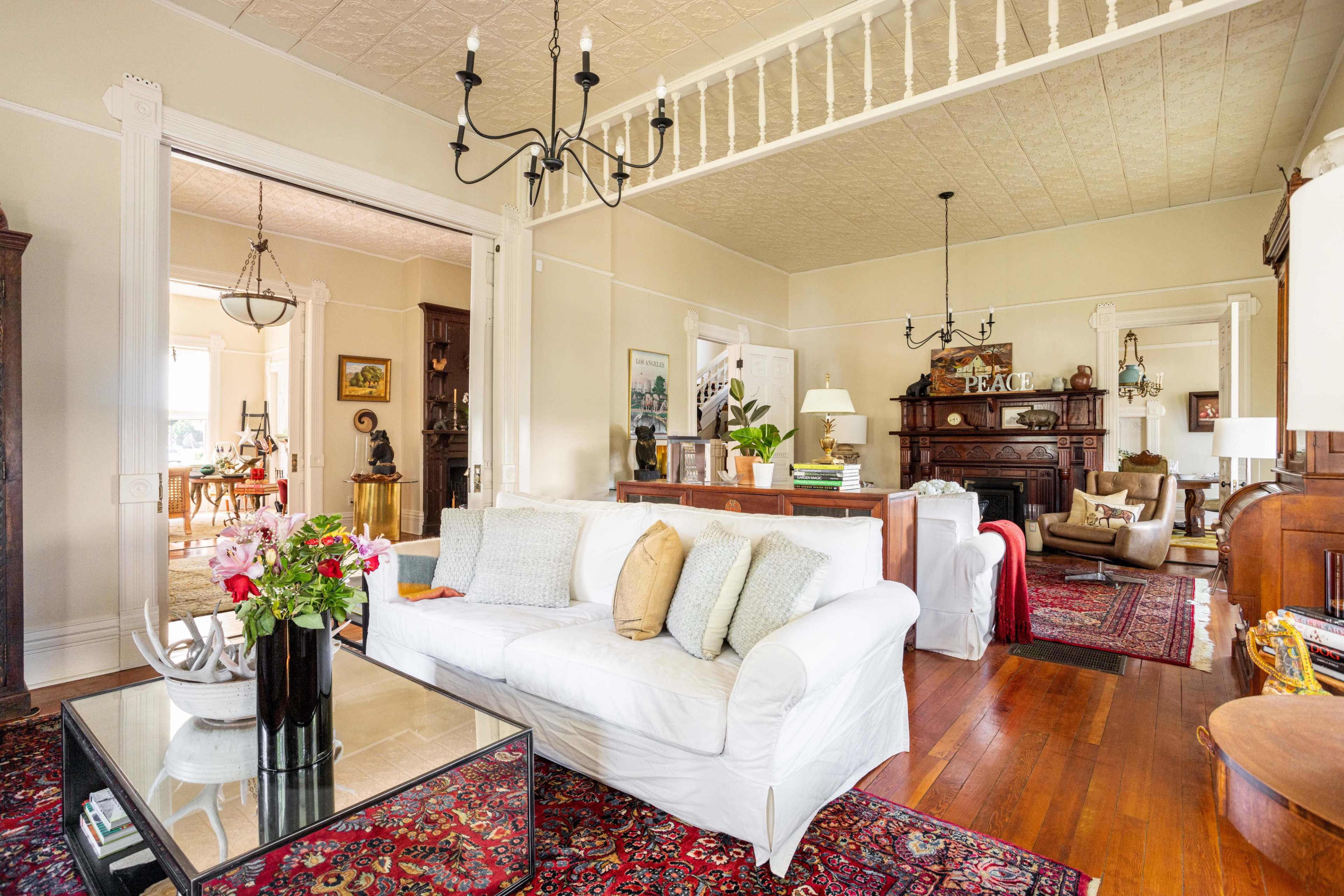 A spacious living room with white furniture, hardwood floors, and ornate decorations, leading to another room visible in the background.